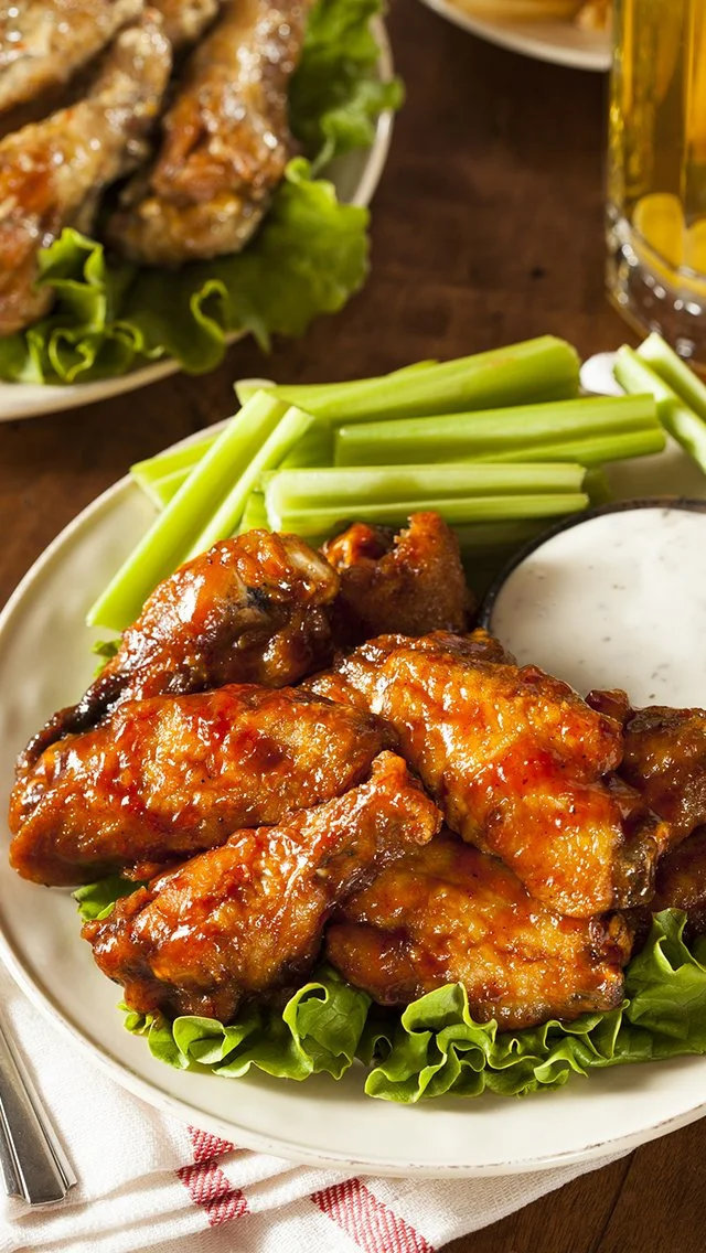 Close-up of buffalo chicken wings with celery sticks and ranch dip on a white plate, with a side of beer in a glass.