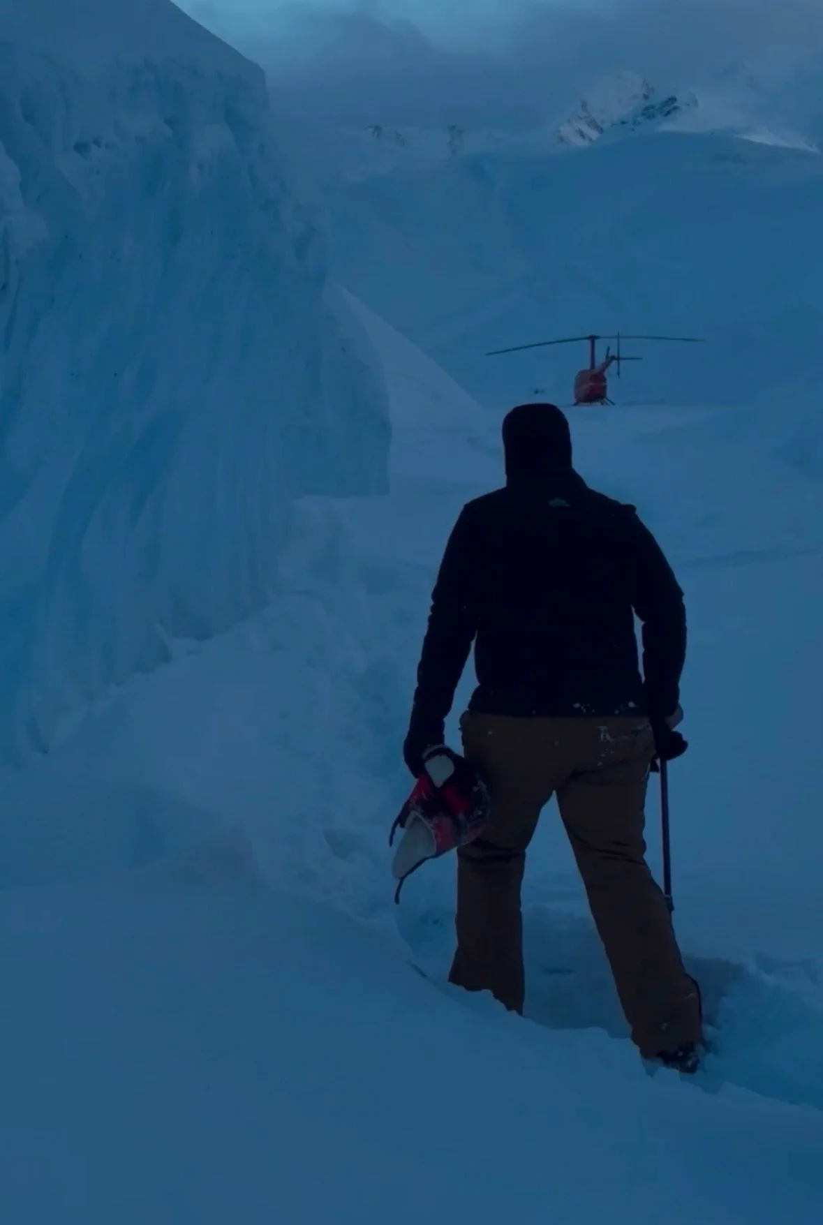 A person standing in a snowy landscape, holding a snow tool in one hand and a helmet, with a helicopter in the background near ice formations.