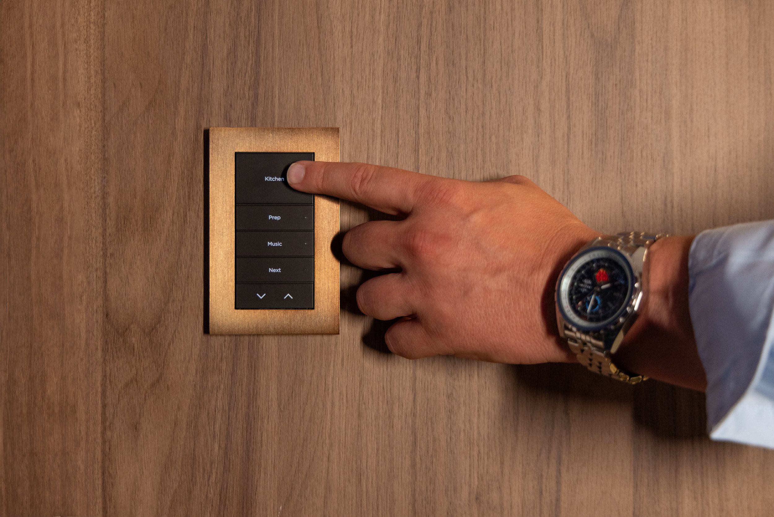 A person pressing a black button labeled 'Kitchen' on a wall-mounted control panel, with other buttons labeled 'Prep,' 'Music,' and 'Next.' The control panel has a gold frame and is installed on a wooden wall. The person's wrist has a silver watch with a black dial.