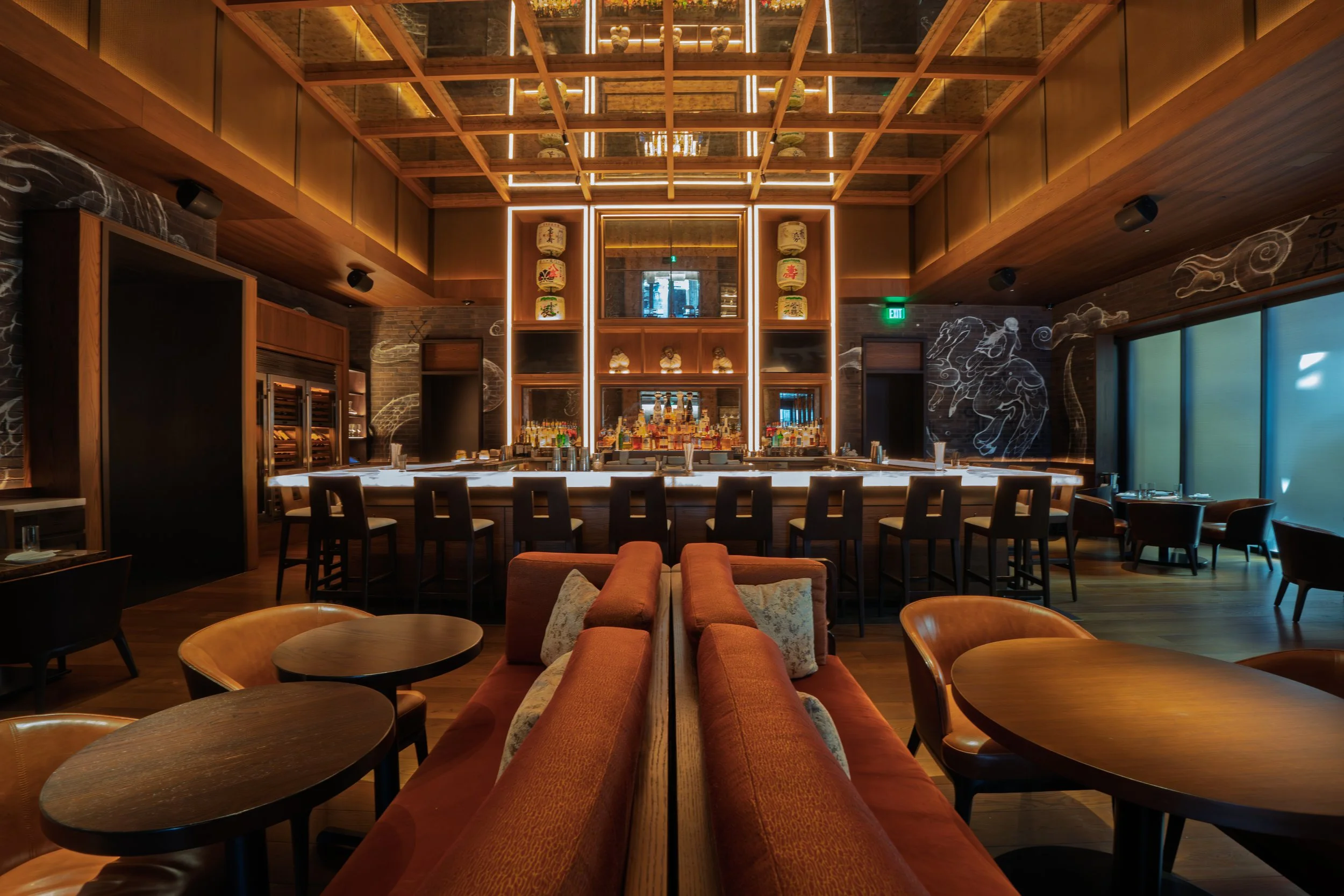 A modern bar and lounge area with a central illuminated bar counter, high chairs, and surrounding tables and chairs, with artistic line drawings of dragons on the walls.