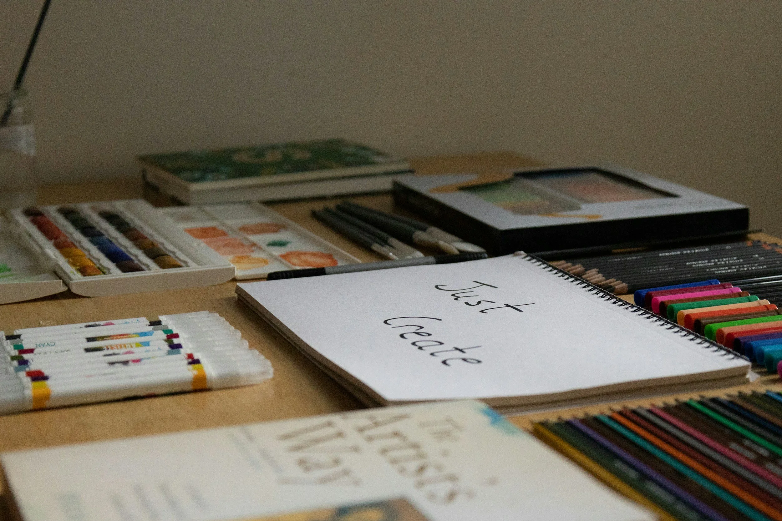 Art supplies including watercolor paints, colored pencils, markers, and a sketchbook with the words 'Just create' written on it are arranged on a wooden table.