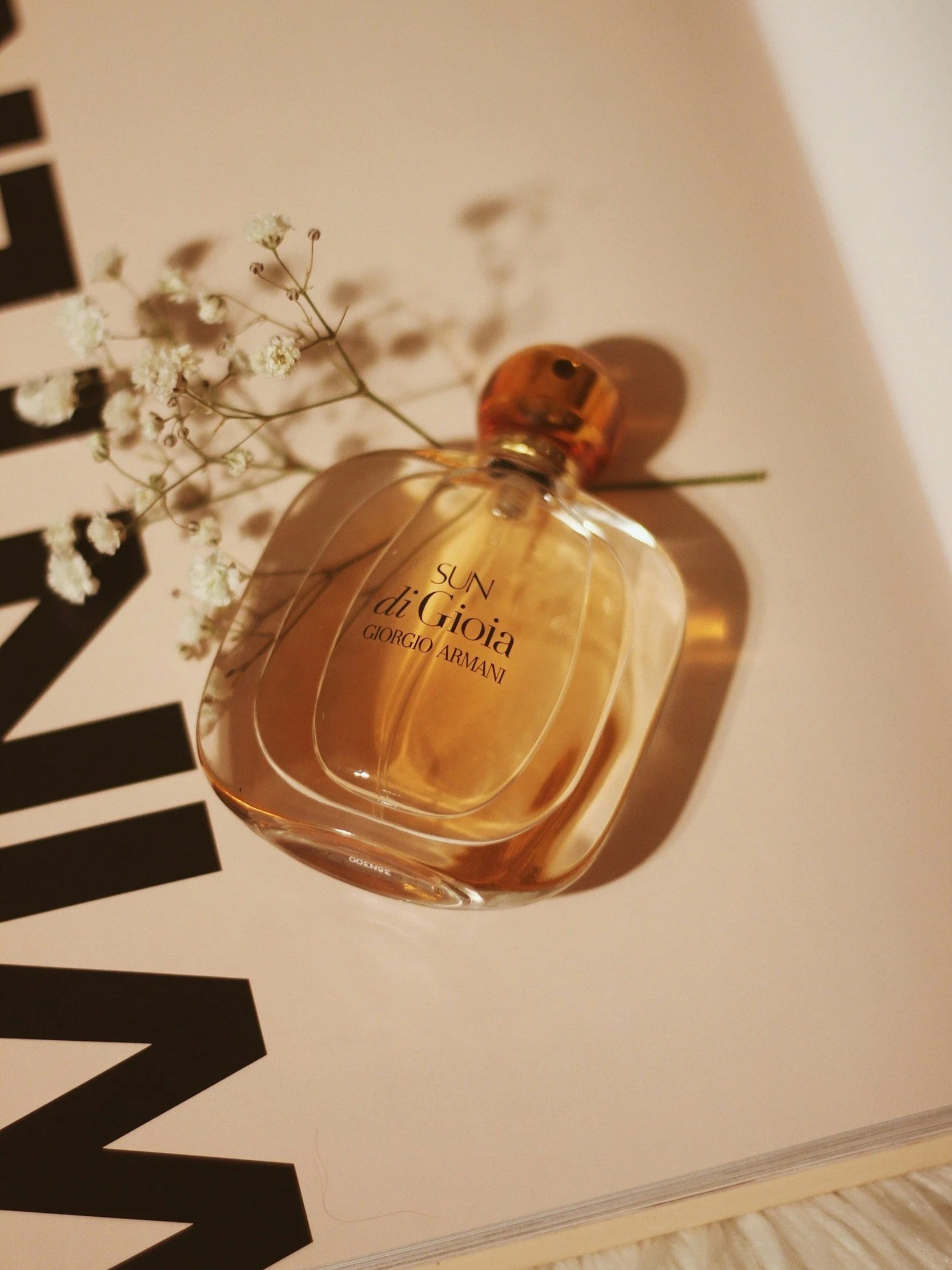A bottle of Giorgio Armani Sun di Gioia perfume resting on a magazine page with black text and a white background, along with small white flowers.