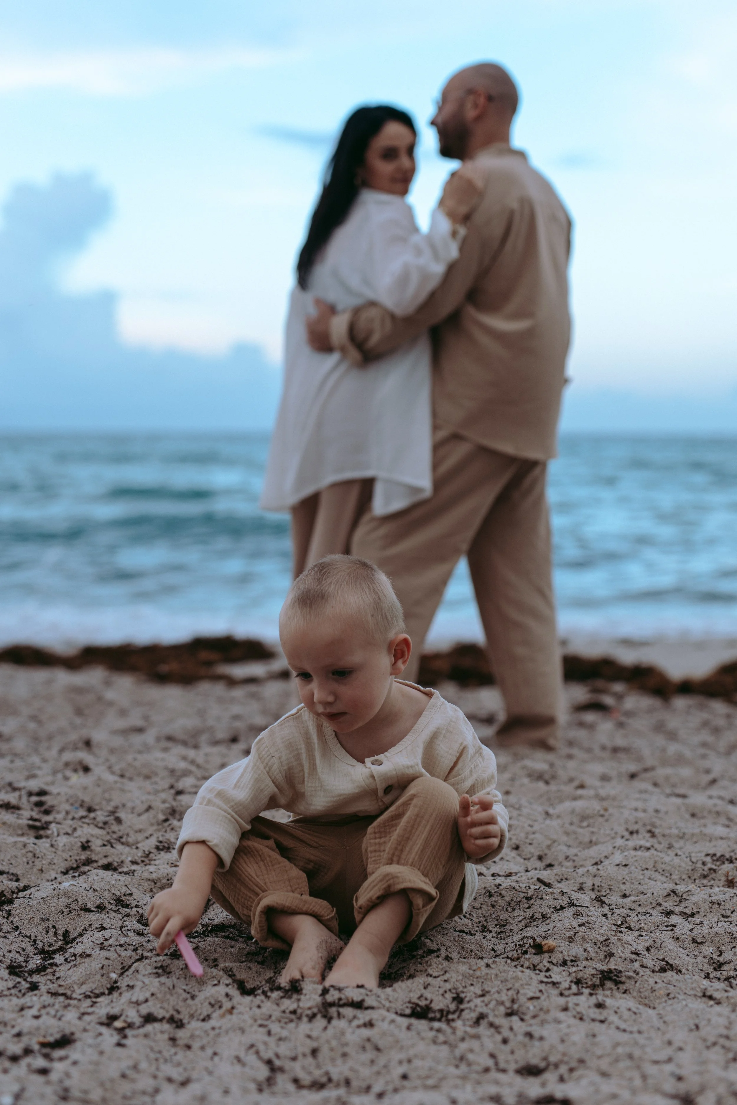 A young boy playing with a toy on the sandy beach, with an ocean and a partly cloudy sky in the background. Behind him, a couple is dancing or embracing by the water, with the woman wearing a white dress and the man in beige clothing.