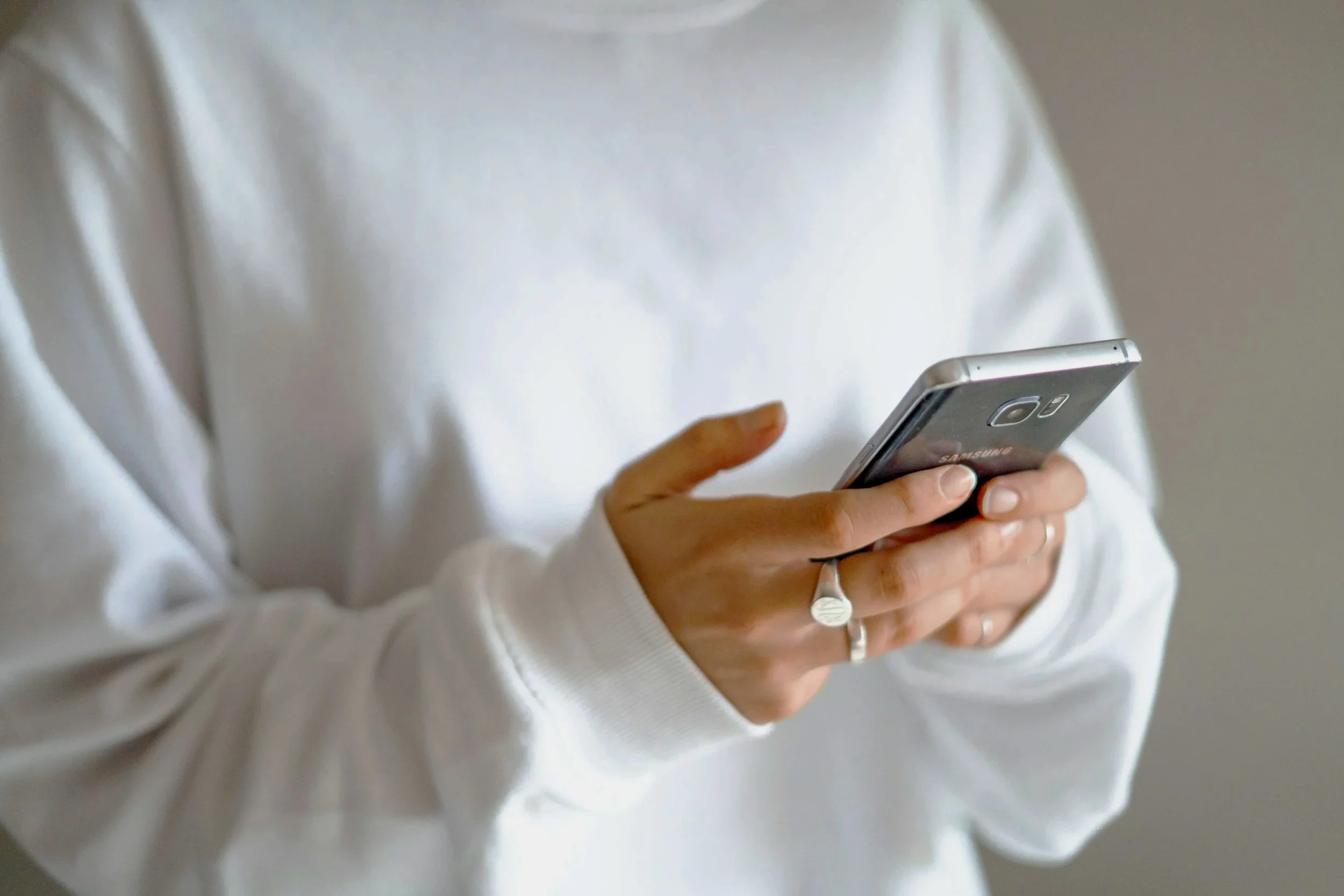 Person holding a silver Samsung smartphone with a ring on their finger, wearing a white long-sleeve shirt.