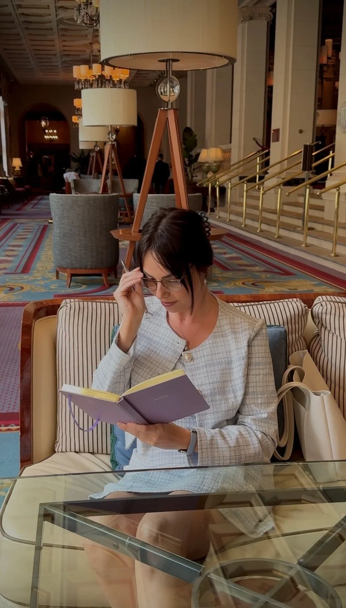 A woman with dark hair, glasses, and a light-colored blazer sitting on a beige and striped sofa in a luxurious hotel lobby, reading a notebook. Behind her are large lamps and elegant interior decor, including a chandelier, staircase, and patterned carpet.