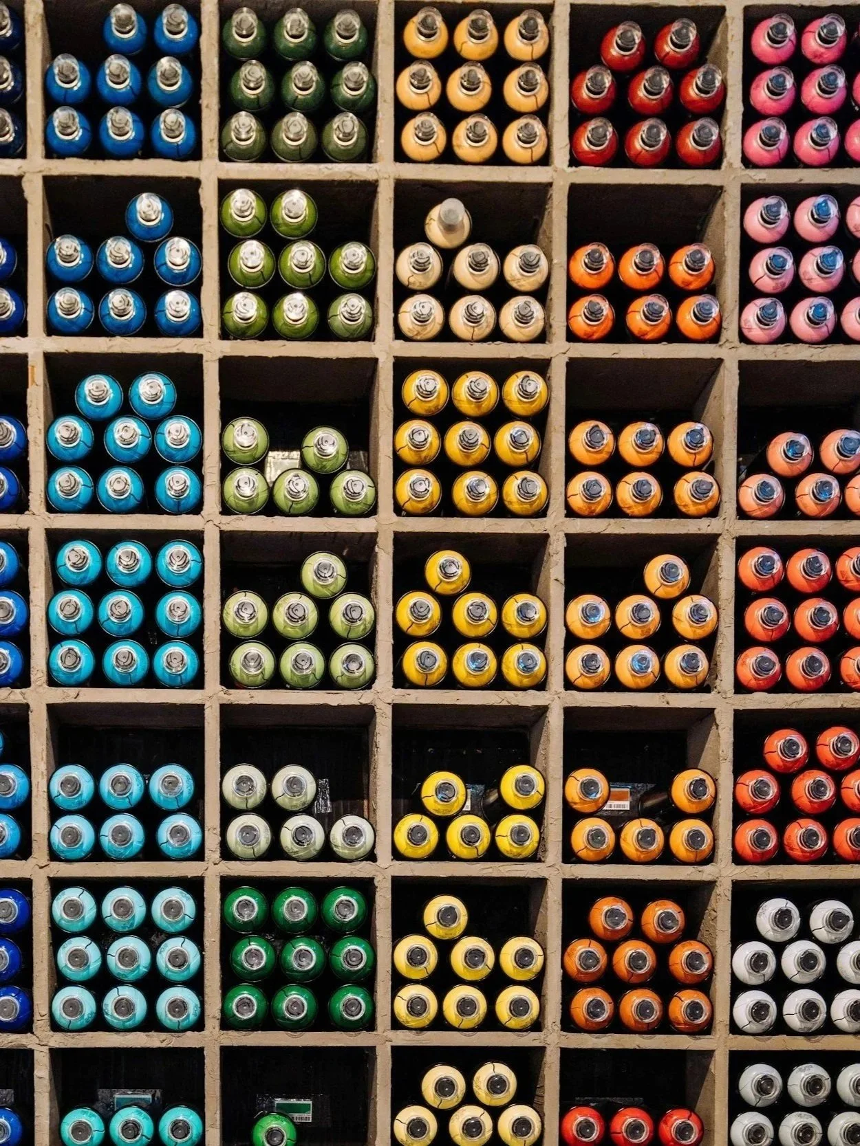 A colorful display of spray paint cans arranged in small square compartments, organized by color in a grid. The colors include blue, green, yellow, orange, pink, and red.