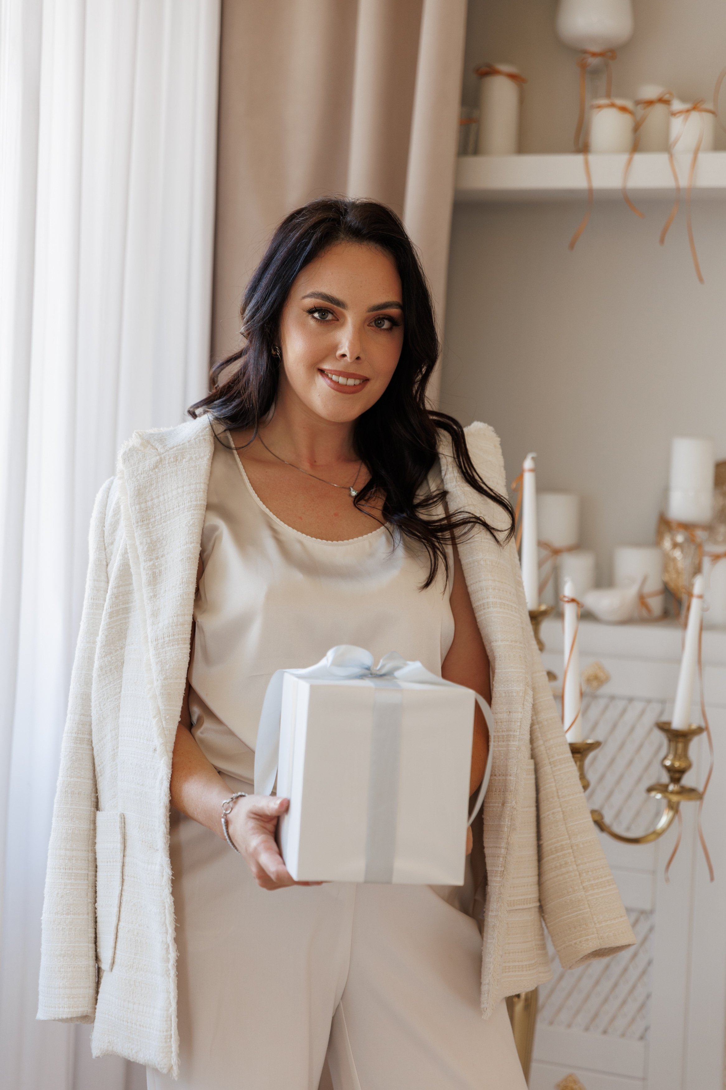 A woman with dark hair wearing a beige dress and a light-colored cardigan, holding a white gift box with a bow, standing indoors with candles and decorative items in the background.