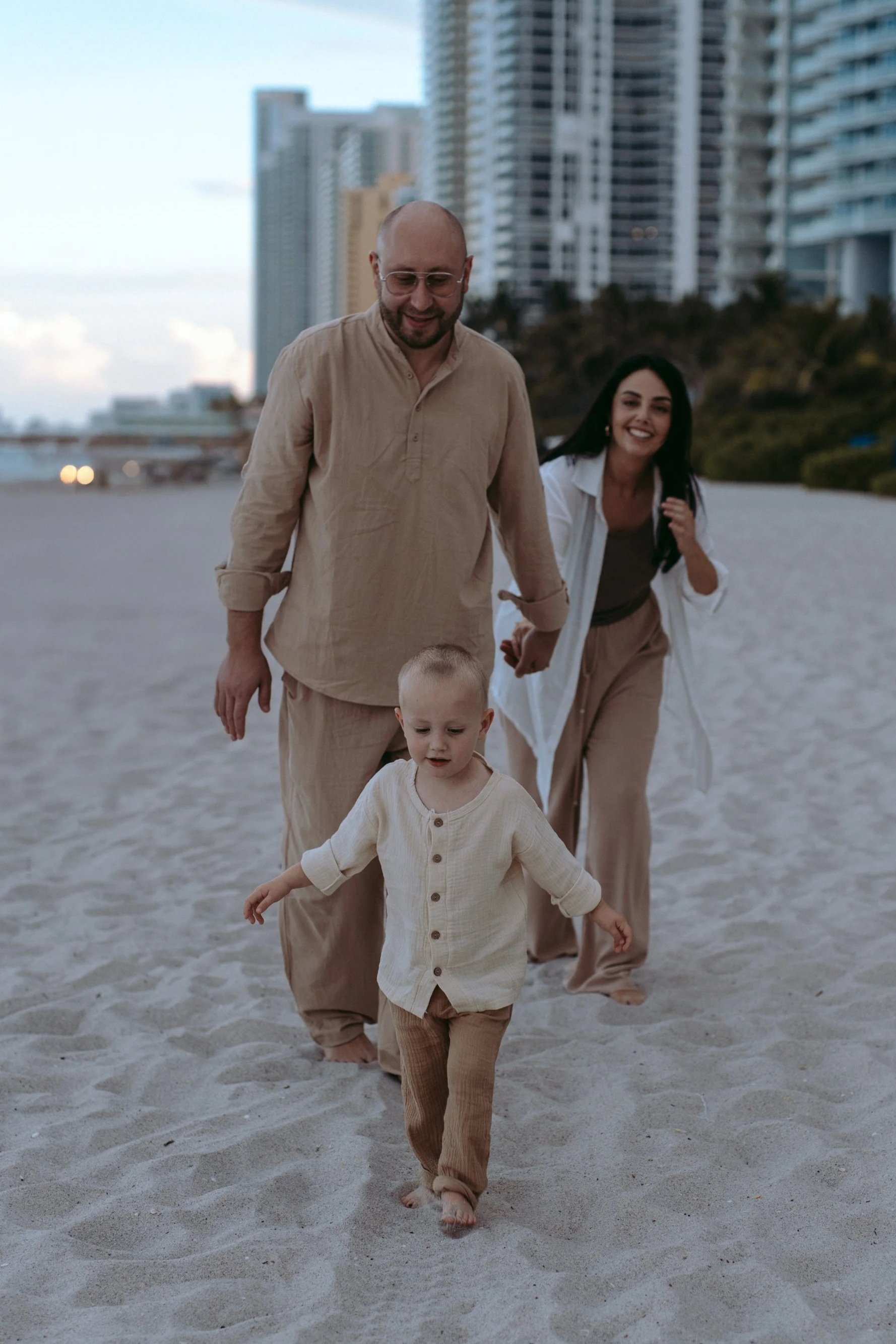 A family walking barefoot on the beach with high-rise buildings in the background during evening time.