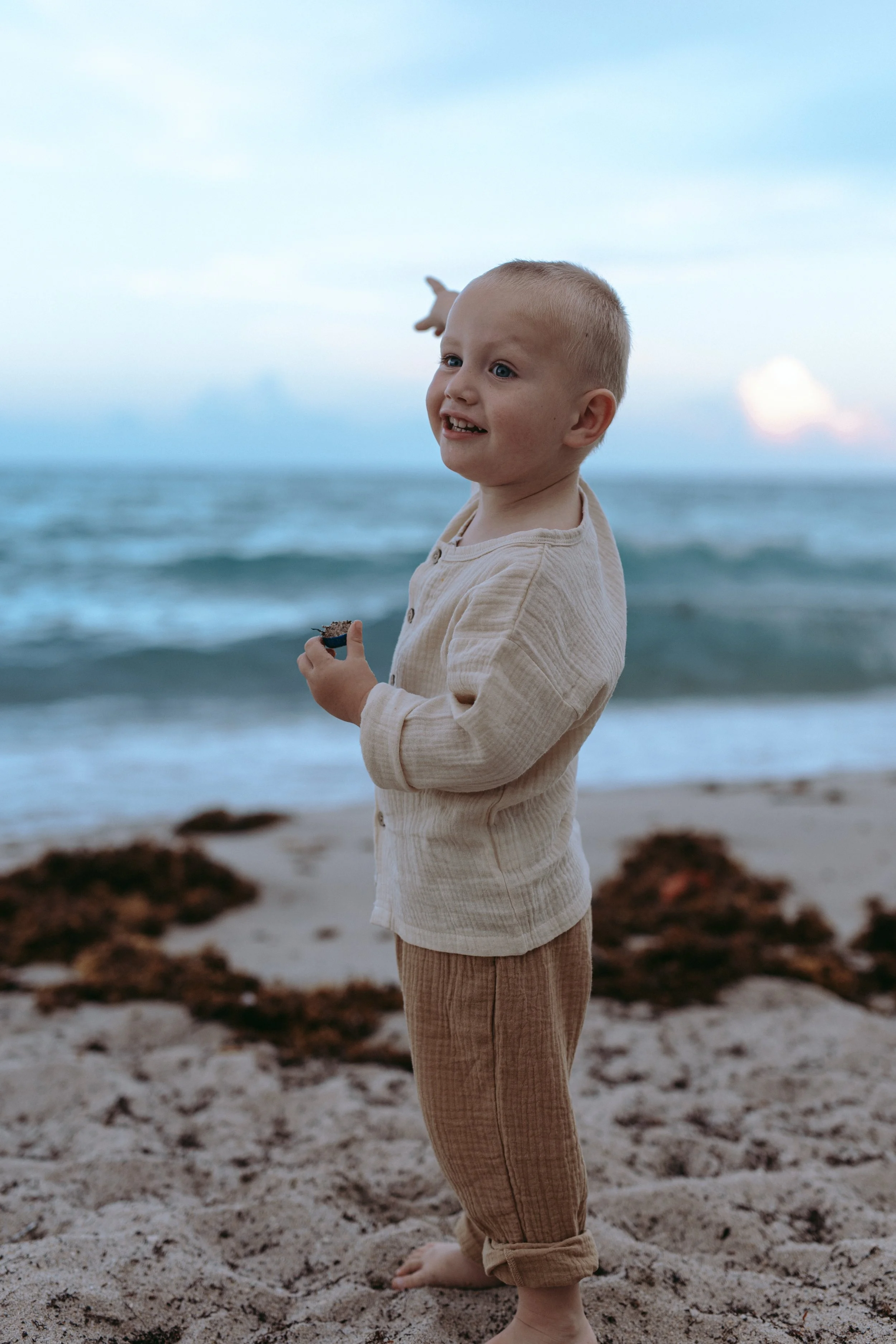 A young boy with blonde hair and fair skin standing barefoot on a sandy beach, looking into the distance with one arm extended, holding a small object, with the ocean and sky in the background.