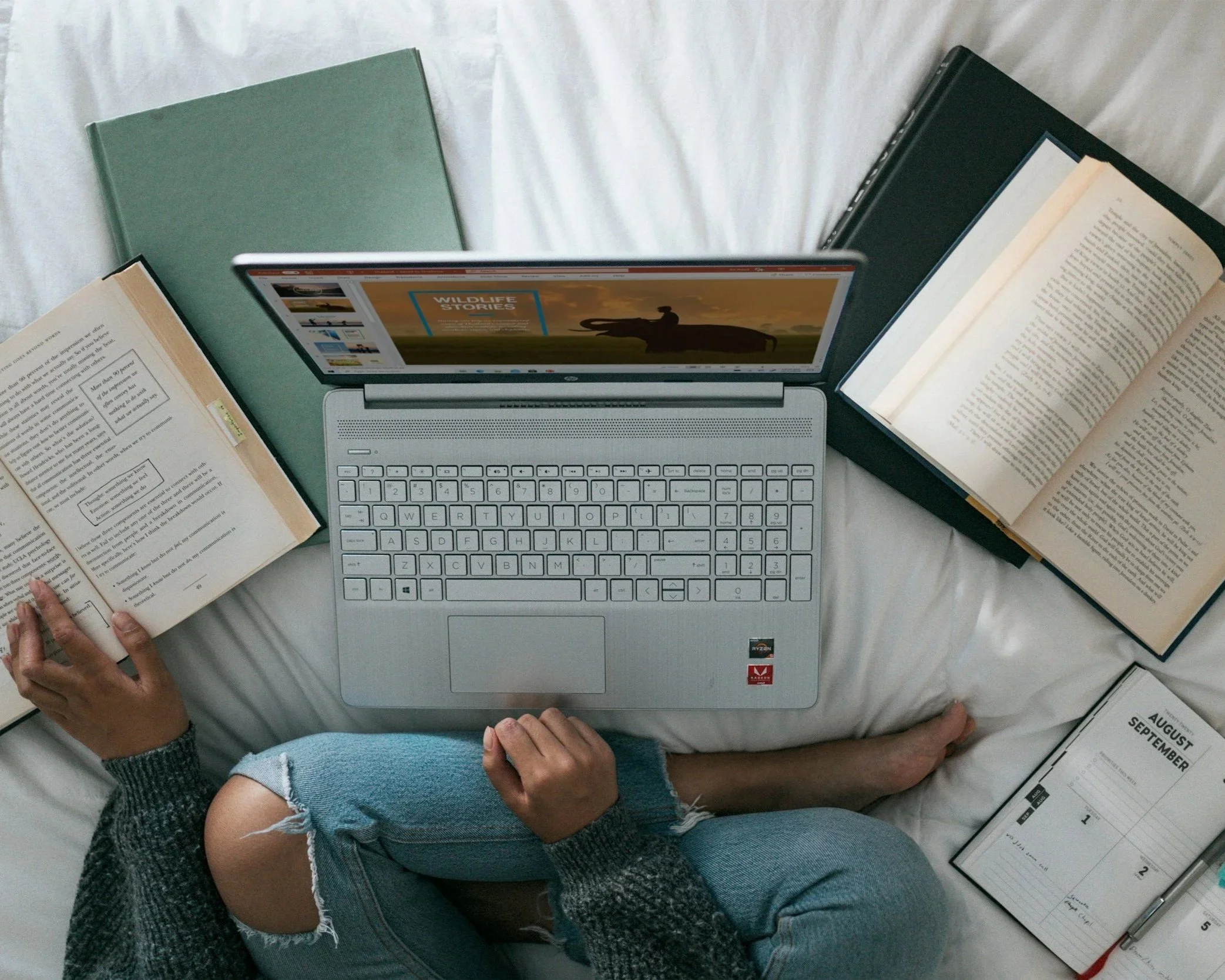 A laptop surrounded by open books on a bed, with hands and arms visible of two people, one in a denim sleeve and another in a gray sweater.