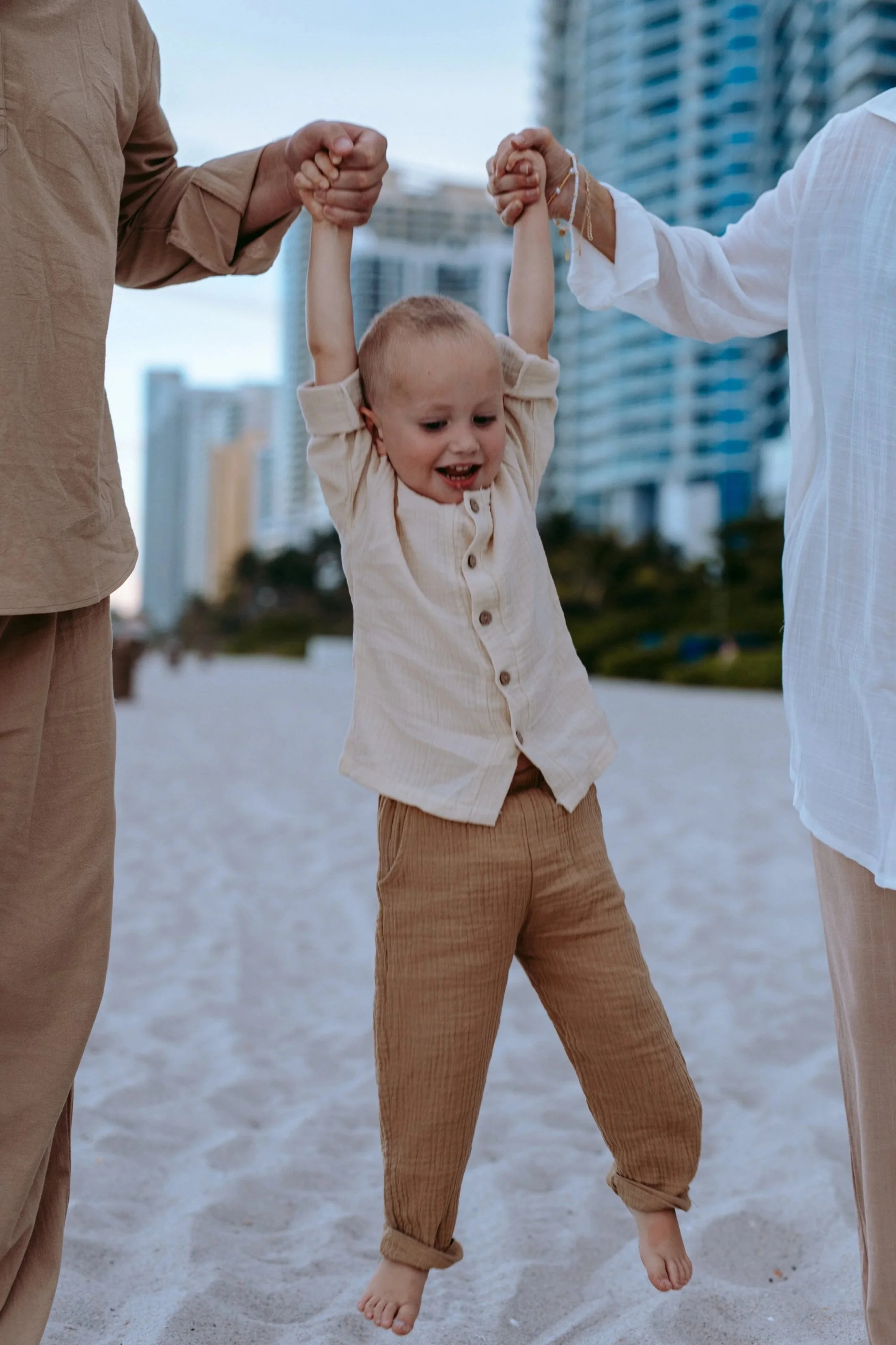 Child being held up by two adults, one on each side, in an outdoor urban setting with tall buildings in the background.
