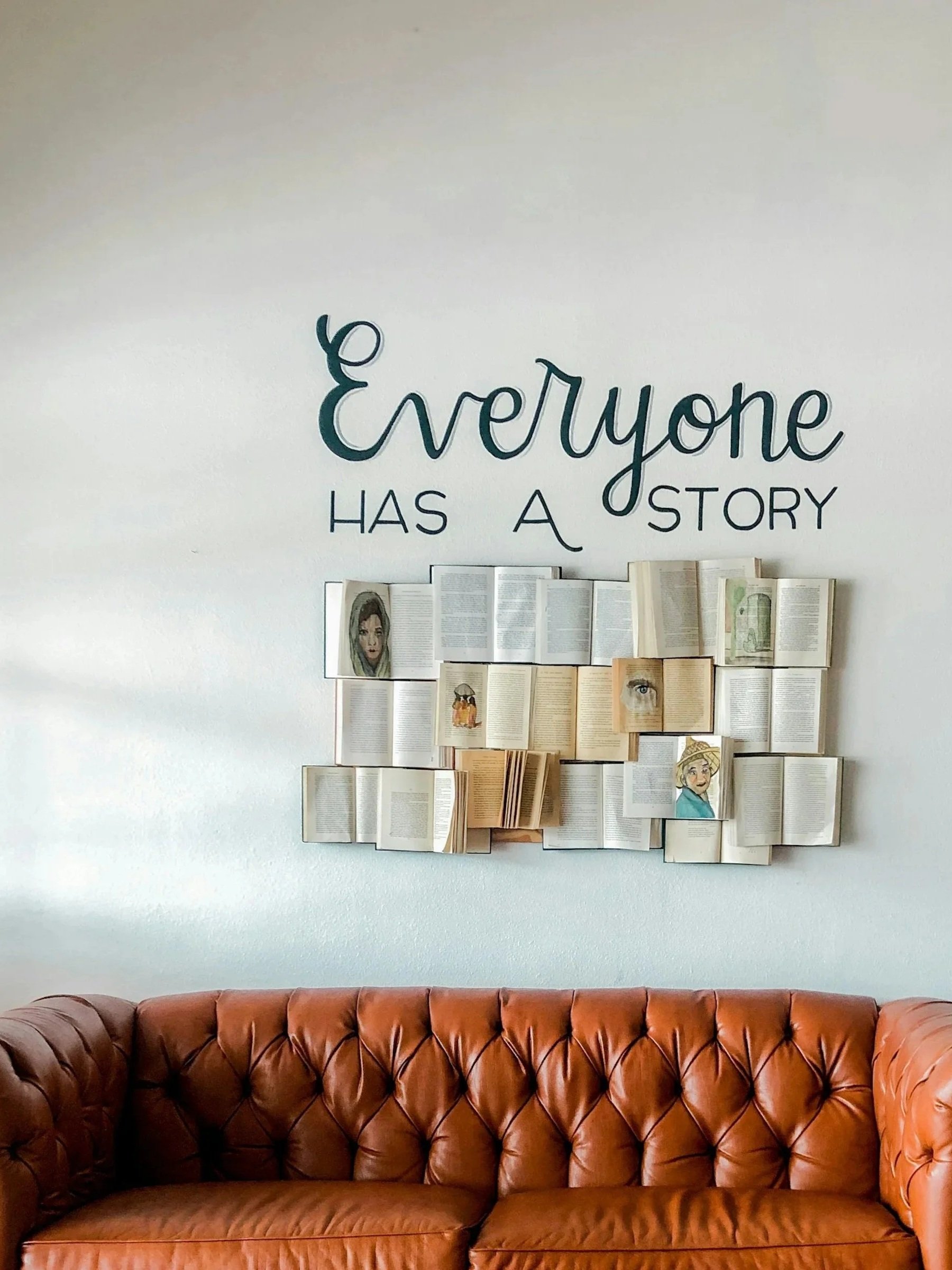 A tan leather sofa in front of a white wall with a decorative display of open books. Above the books, a large wall decal reads 'Everyone has a story' in blue script font.