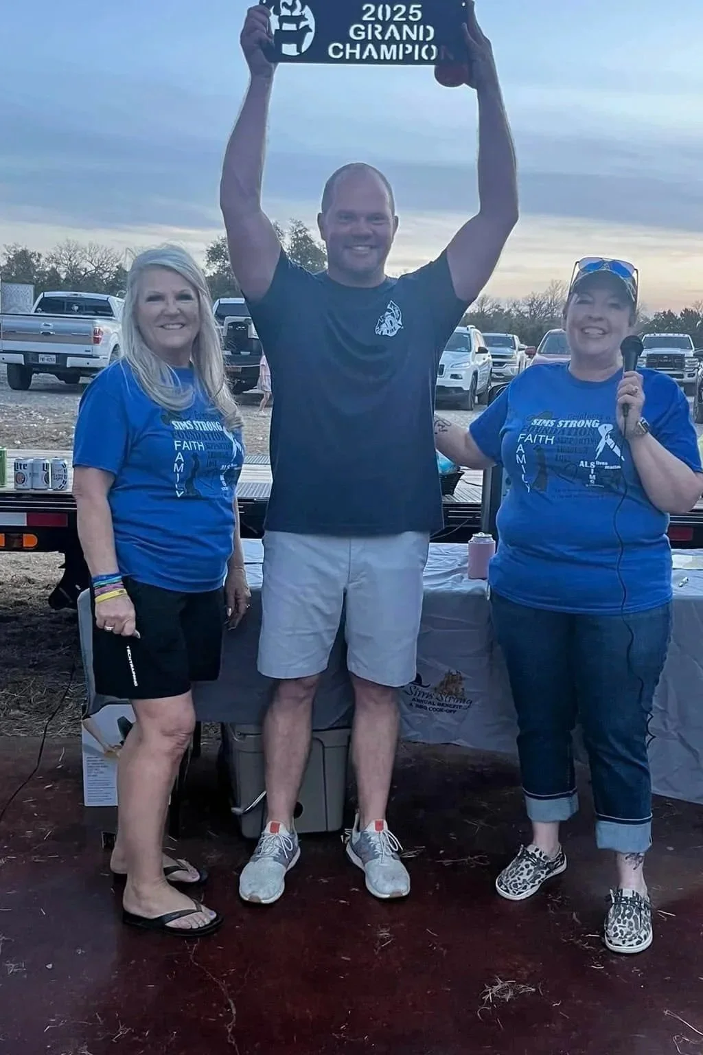 Three people standing outdoors at a fundraising event, with a man in the center holding a sign that reads '2025 GRAND CHAMPION'. The two women on either side are smiling, dressed in matching blue T-shirts with supportive messages. One woman is holdin
