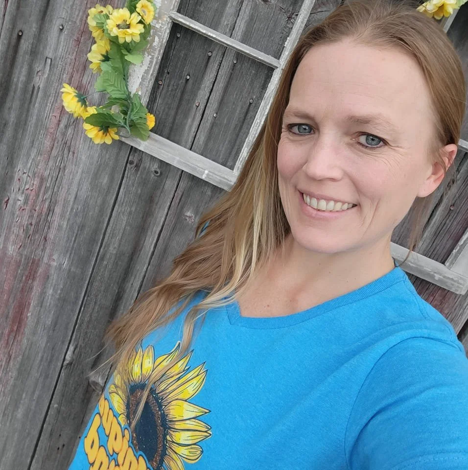 A smiling woman with long blonde hair wearing a blue t-shirt with a sunflower graphic, taking a selfie outdoors with a weathered wooden fence and yellow sunflower decoration in the background.