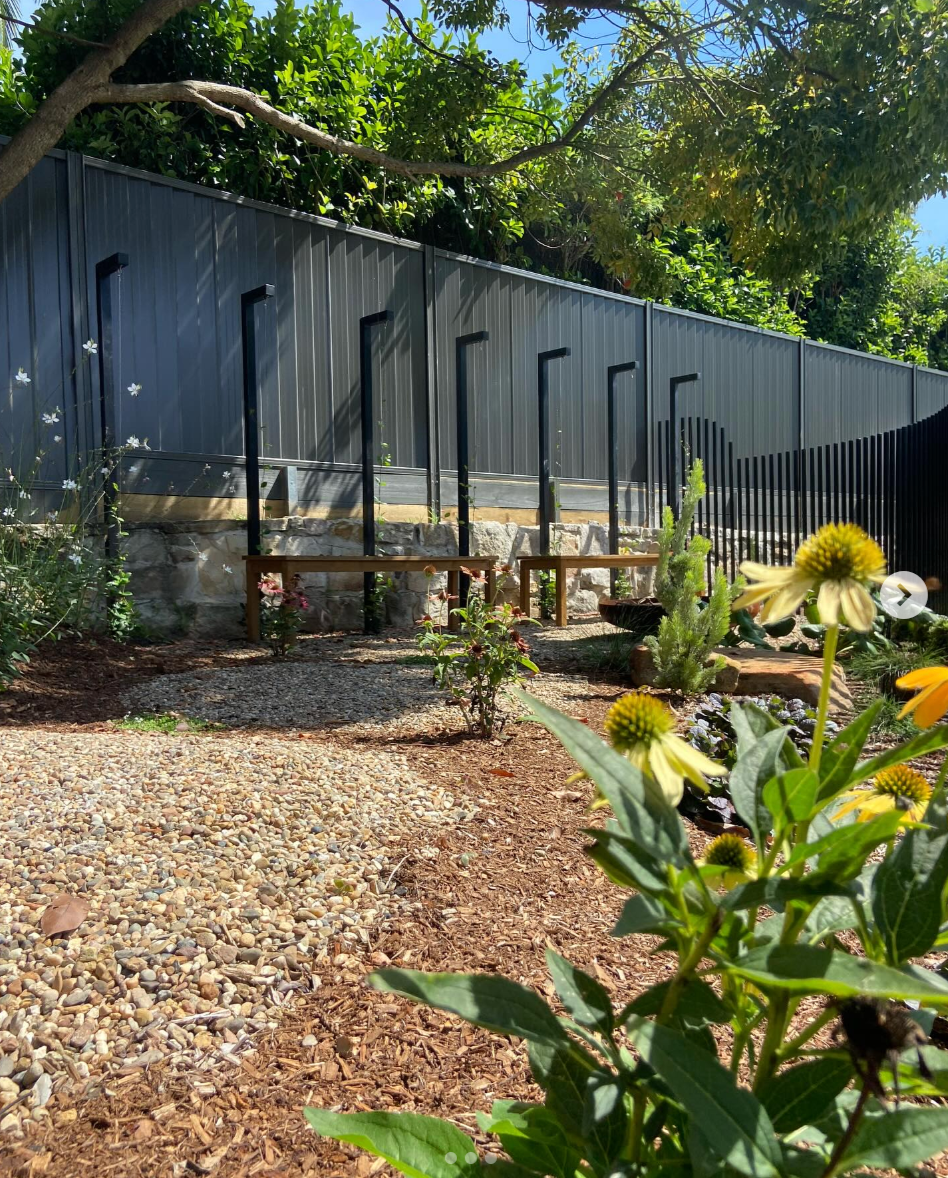 A garden with flowering plants, a gravel and dirt path, a black metal fence, and a blue sky with some green trees overhead.