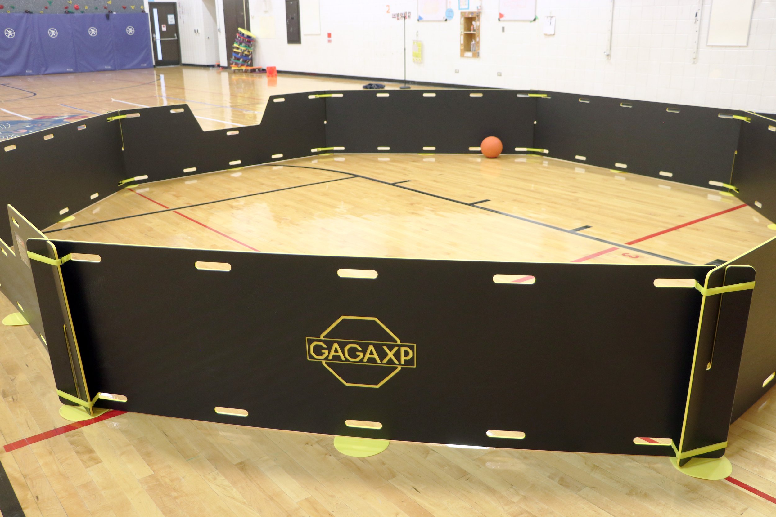 Indoor gymnasium with black gaga pit labeled GAGAXP and a small orange ball inside the barrier.