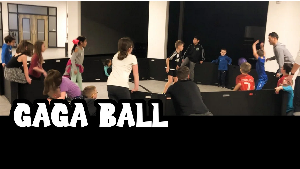 Children and adults playing Gaga Ball inside an indoor arena with padded walls.