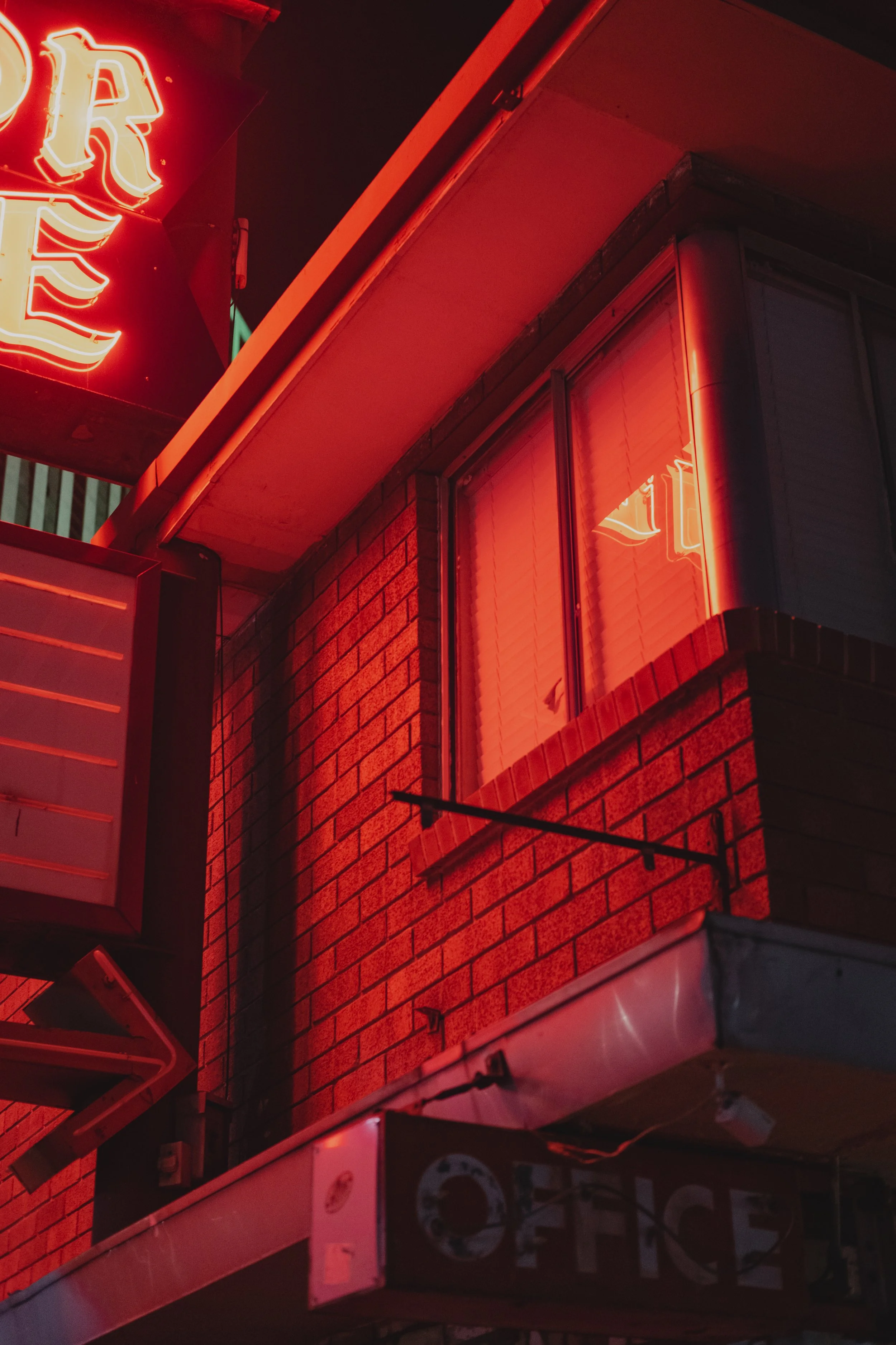 Neon red lights illuminating the exterior of a building, highlighting a portion of the word 'RES' on a sign, with reflections in a window above an office sign.