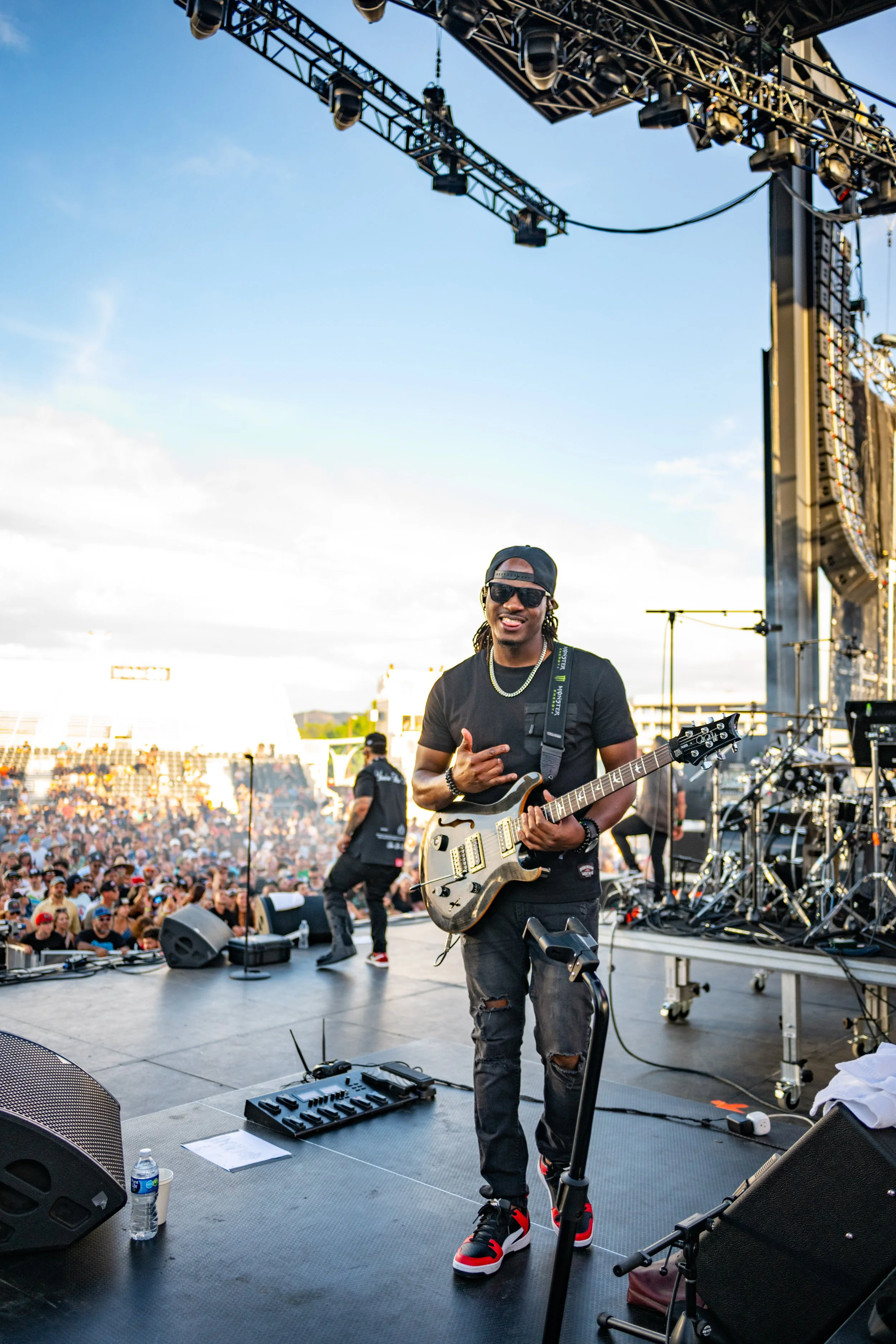 A musician on stage holding an electric guitar, smiling, wearing sunglasses, a black cap backwards, black t-shirt, ripped jeans, headphones around his neck, and a chain, with a large crowd in the background at an outdoor concert during daylight.