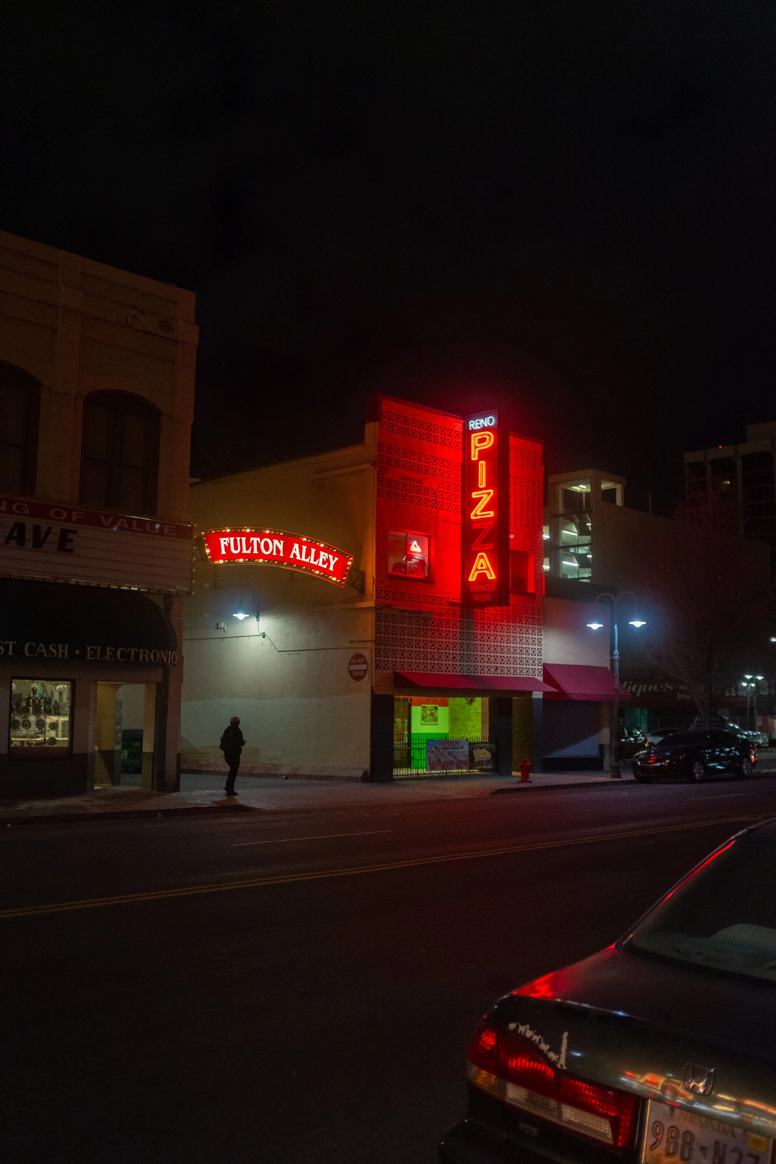 Night view of a restaurant with a bright red neon sign that says 'PIZZA' and a smaller sign above it that says 'RENO.' To the left, there is a marquee sign with the words 'FULTON ALLEY.' The scene includes a pedestrian walking on the sidewalk and parked cars along the street.