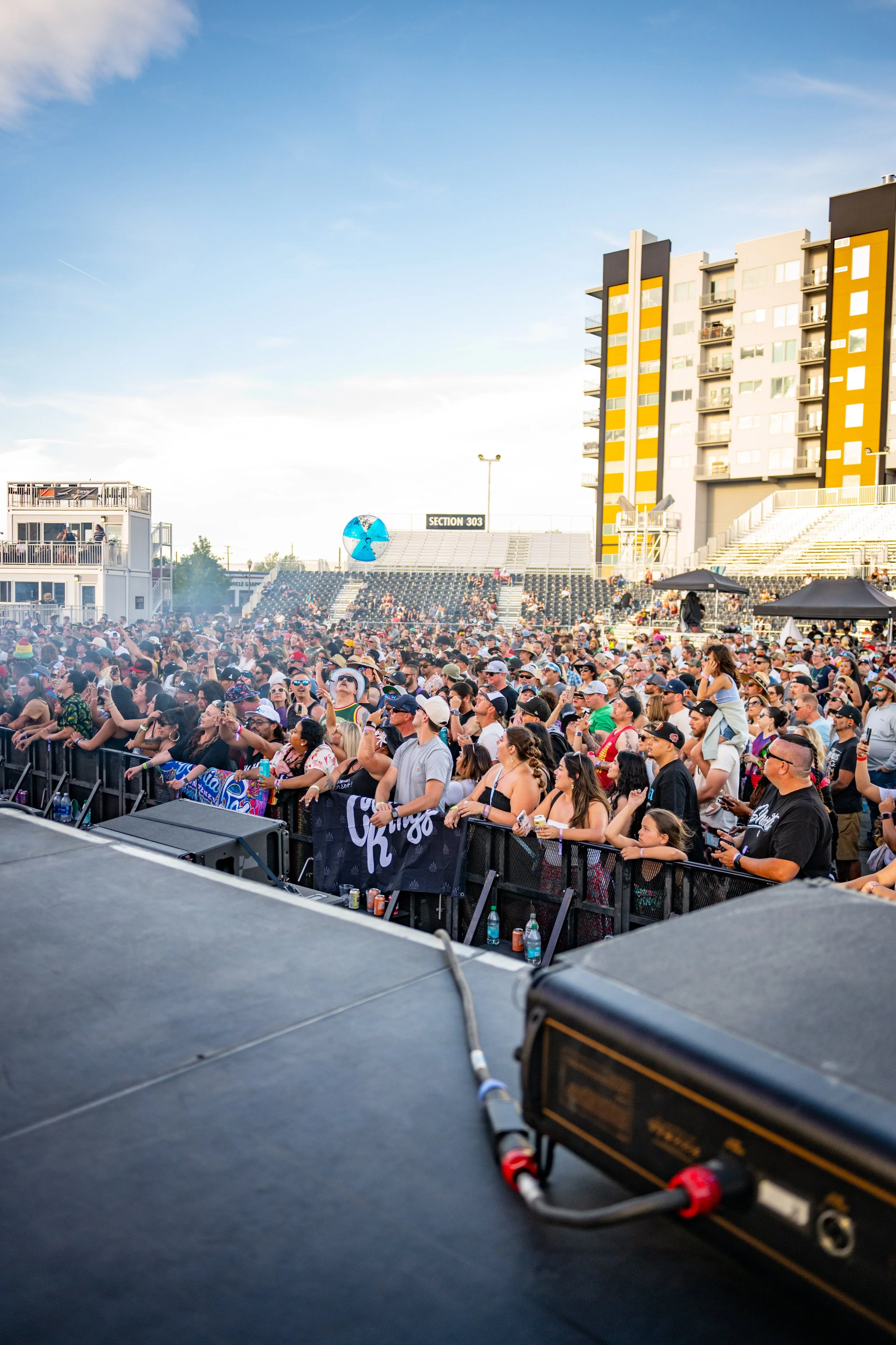 Crowd of people at a music concert, seen from the stage perspective, with high-rise buildings in the background on a clear day.
