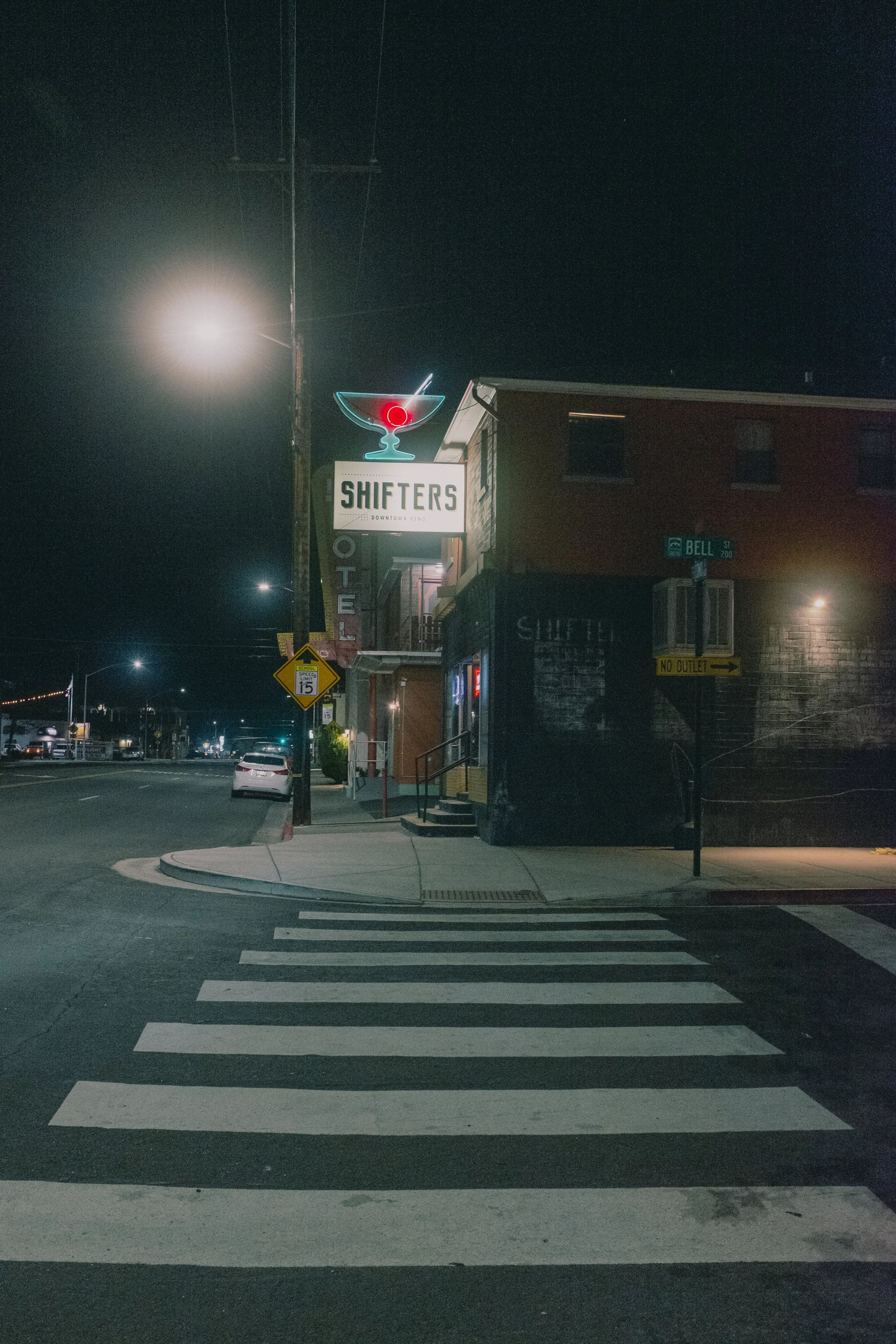 Nighttime street corner with a lit neon sign for 'Shifters' bar, a crosswalk in the foreground, and a building with the bar's sign on the side. Street lamps illuminate the scene, and few cars are seen on the street.
