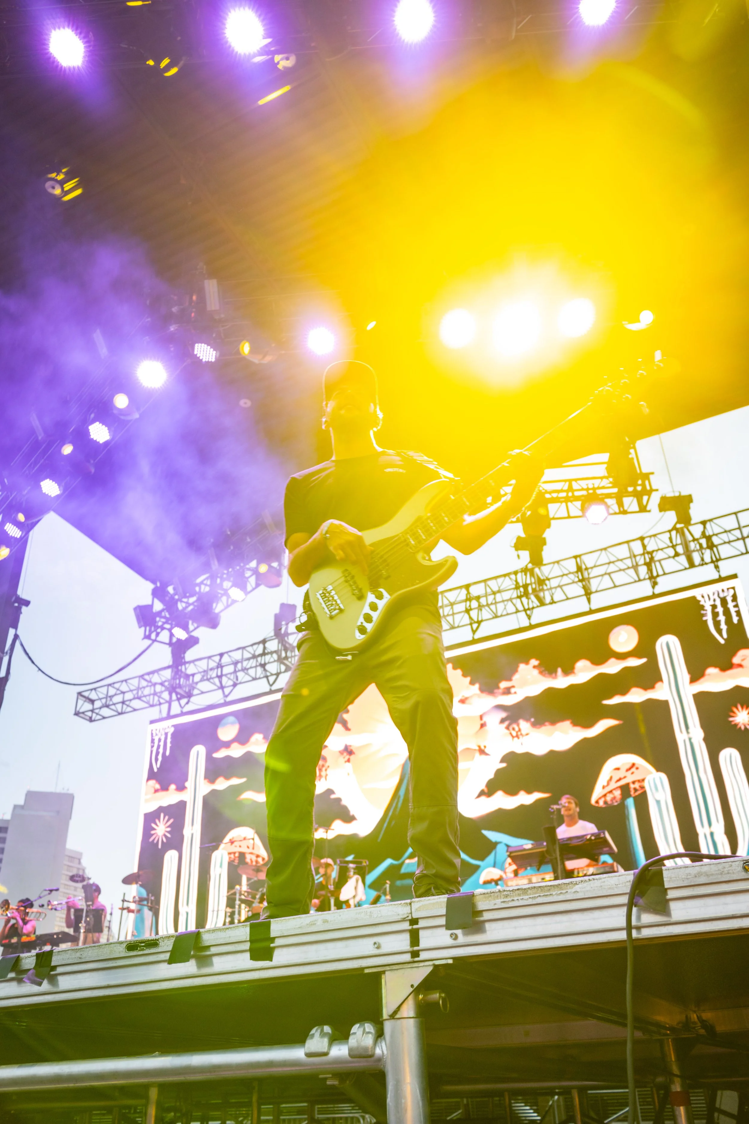 A musician playing an electric guitar on stage during a concert, with bright yellow and purple stage lights overhead and a large screen displaying artwork in the background.