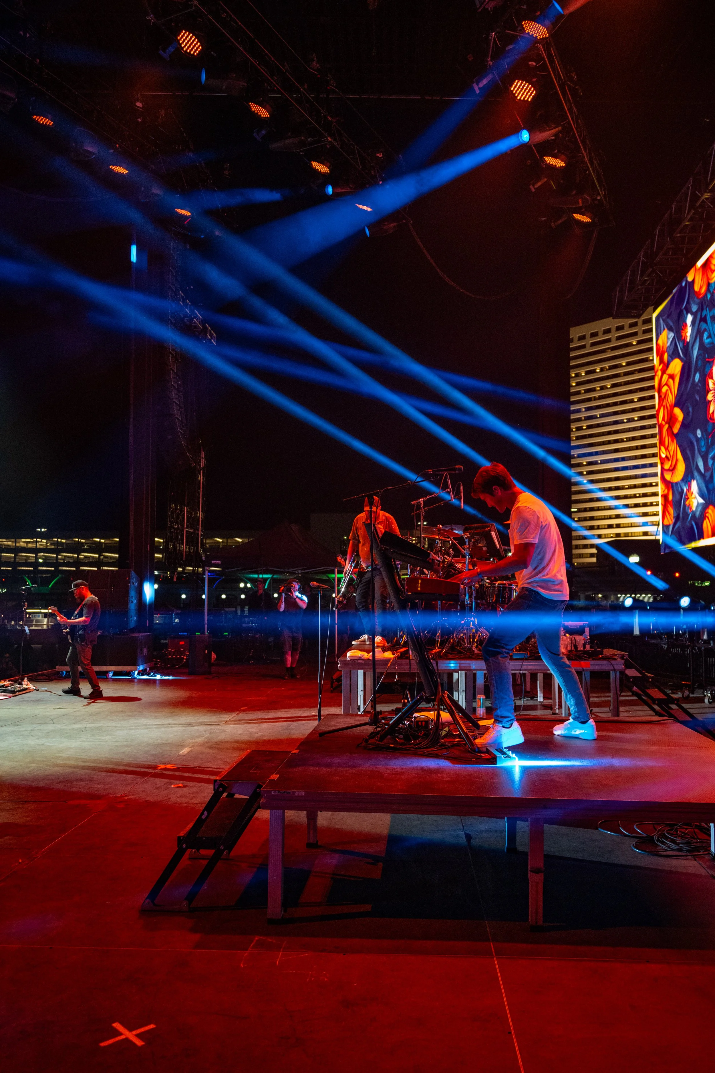 A live musical performance on stage with vibrant blue and orange lights, showing musicians playing instruments, including a keyboardist in the foreground.