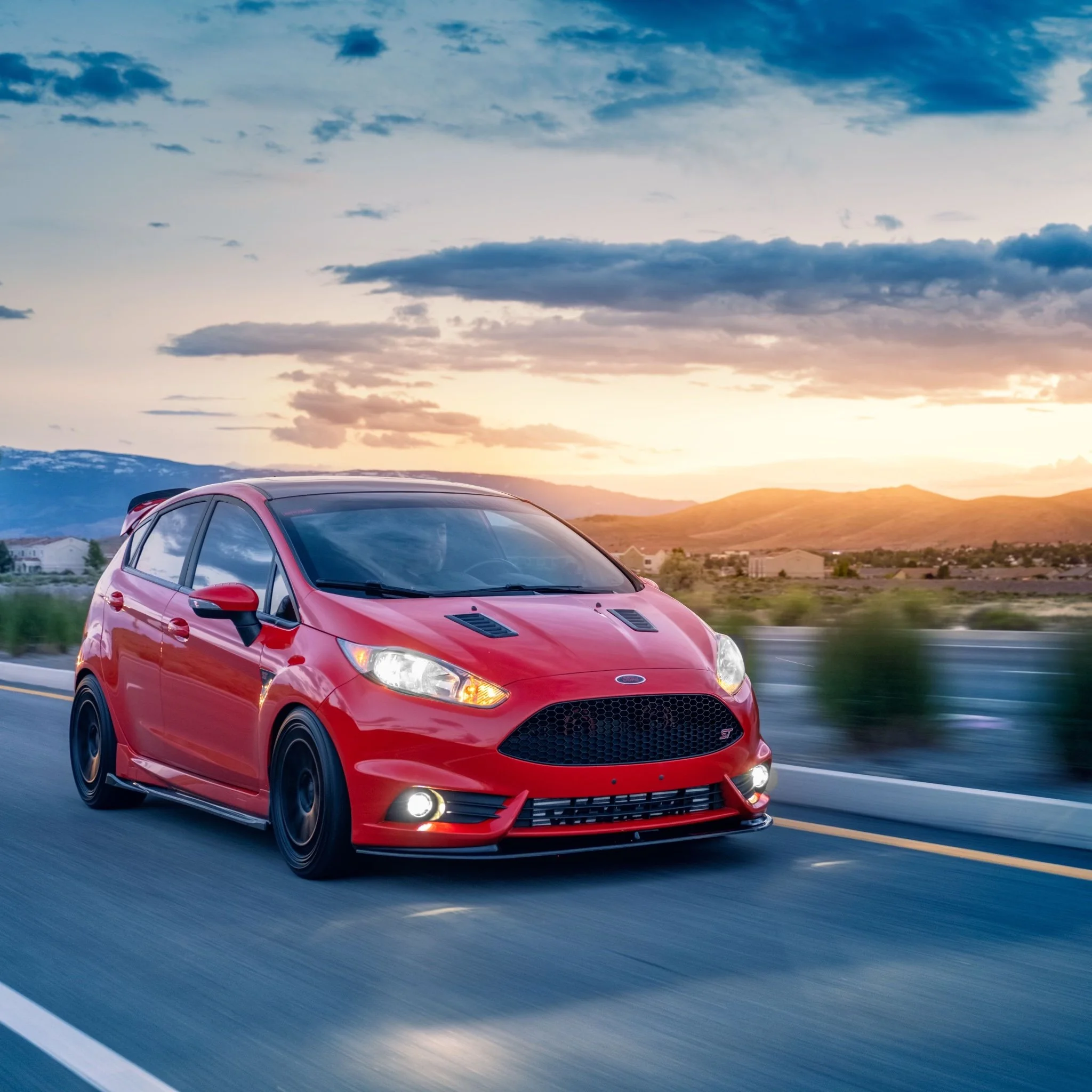 A red Ford Fiesta driving on an open road at sunset in a mountainous area.