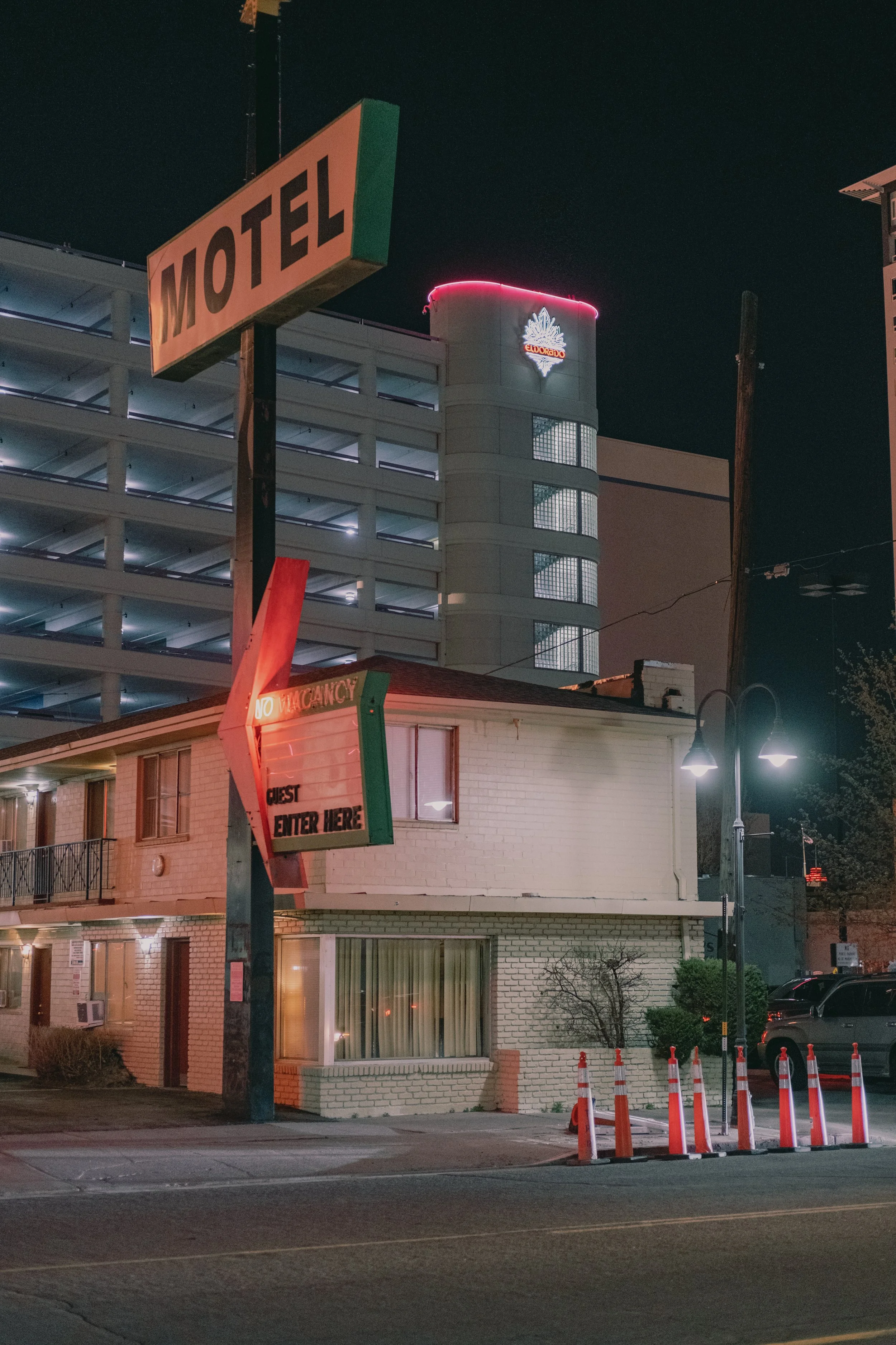Nighttime street scene with a motel sign, a two-story building, parking lot with cars, orange traffic cones, and a tall building with neon lights in the background.