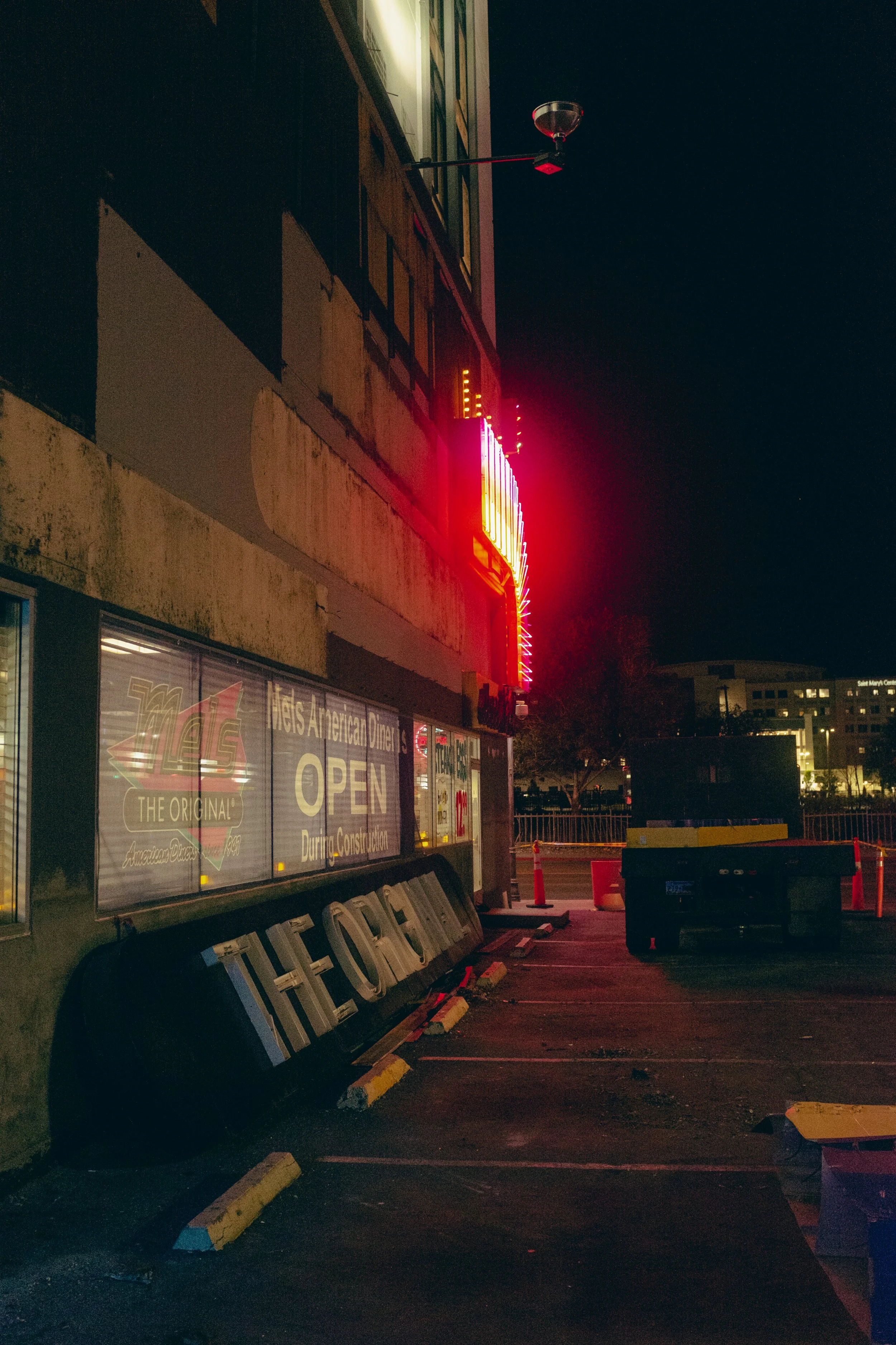 Nighttime scene of a diner with neon lights, a sign reading 'OPEN' in the window, and parking spaces with orange cones and barriers.