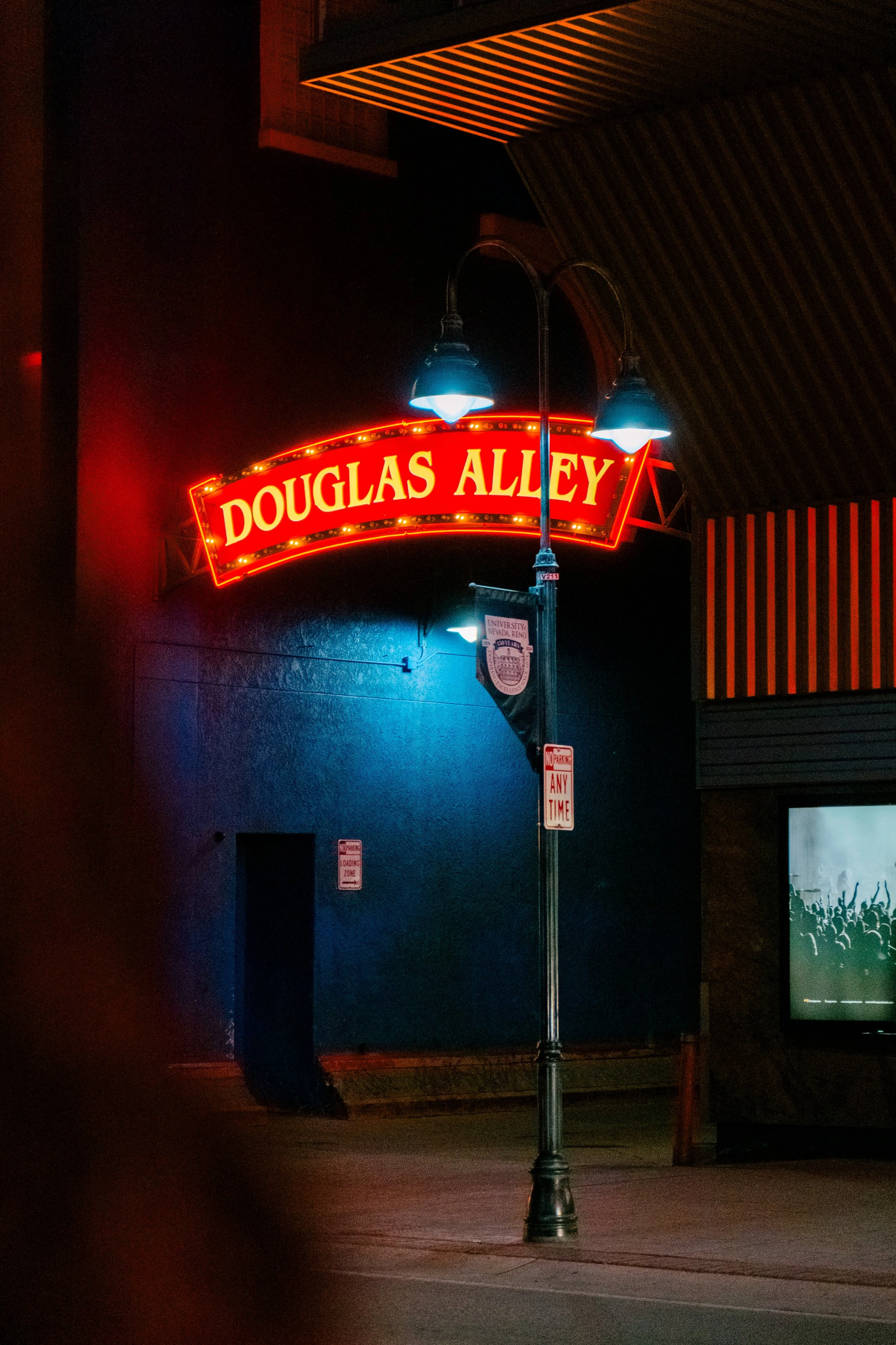 Night scene of a street with a bright red neon sign reading 'DOUGLAS ALLEY,' a lamppost with blue lights, and a blue wall in the background.