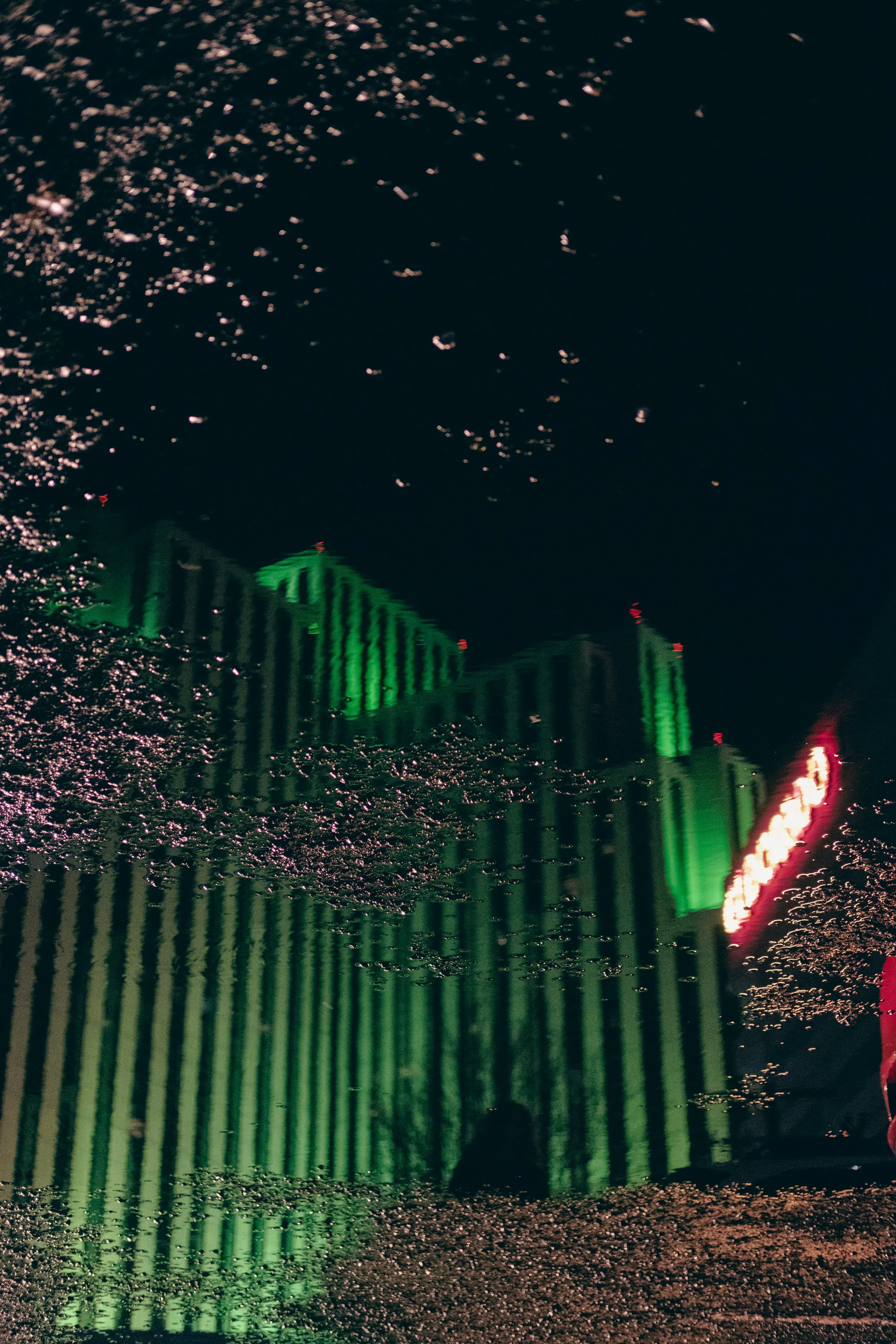 Reflected image of a tall building with green lighting in a puddle, with pinkish sparkles from light reflections and some red and white lights on the right side.