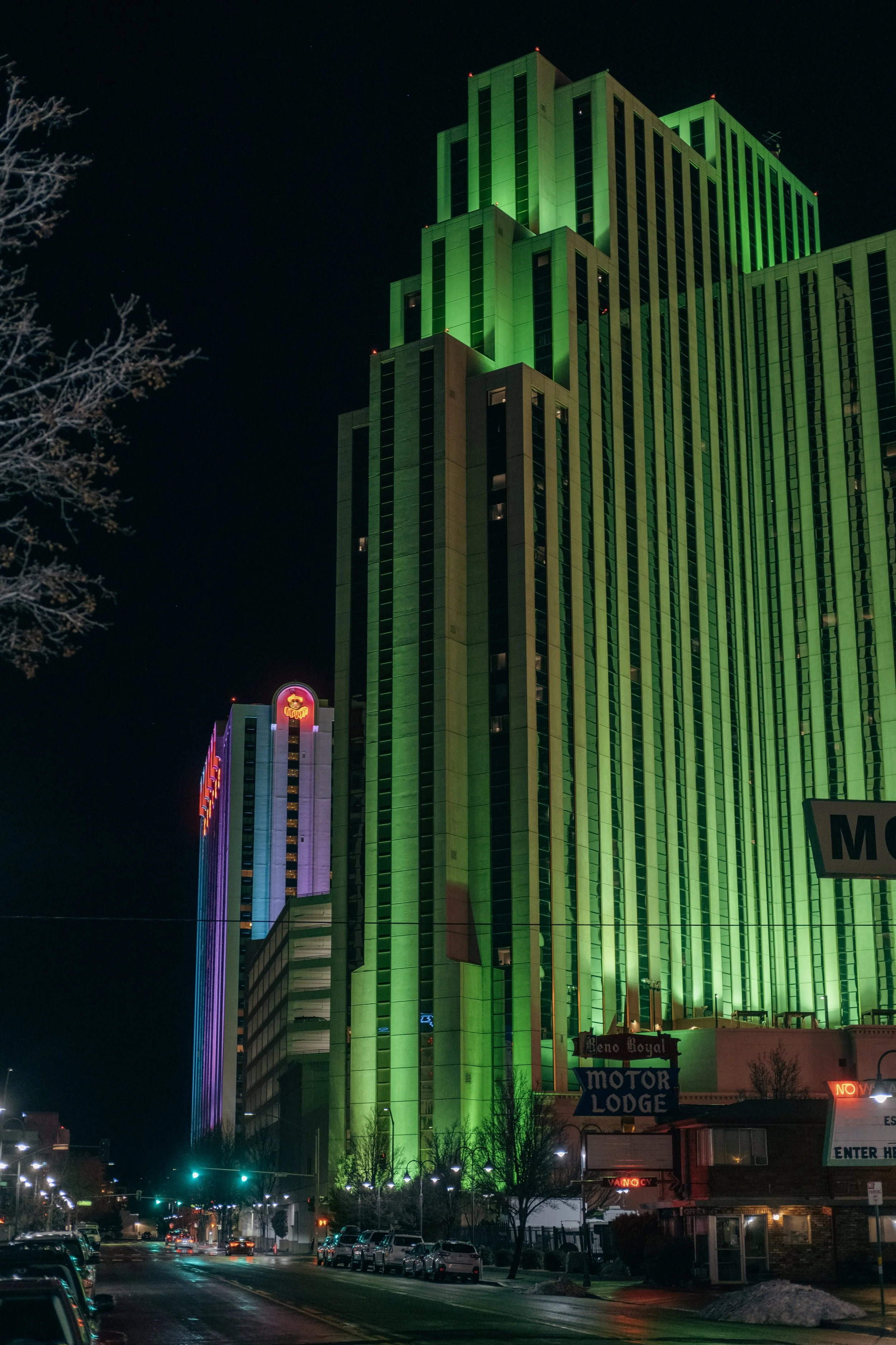 Night view of a tall, high-rise hotel building illuminated in green lights, with smaller buildings and parking lot in the foreground.