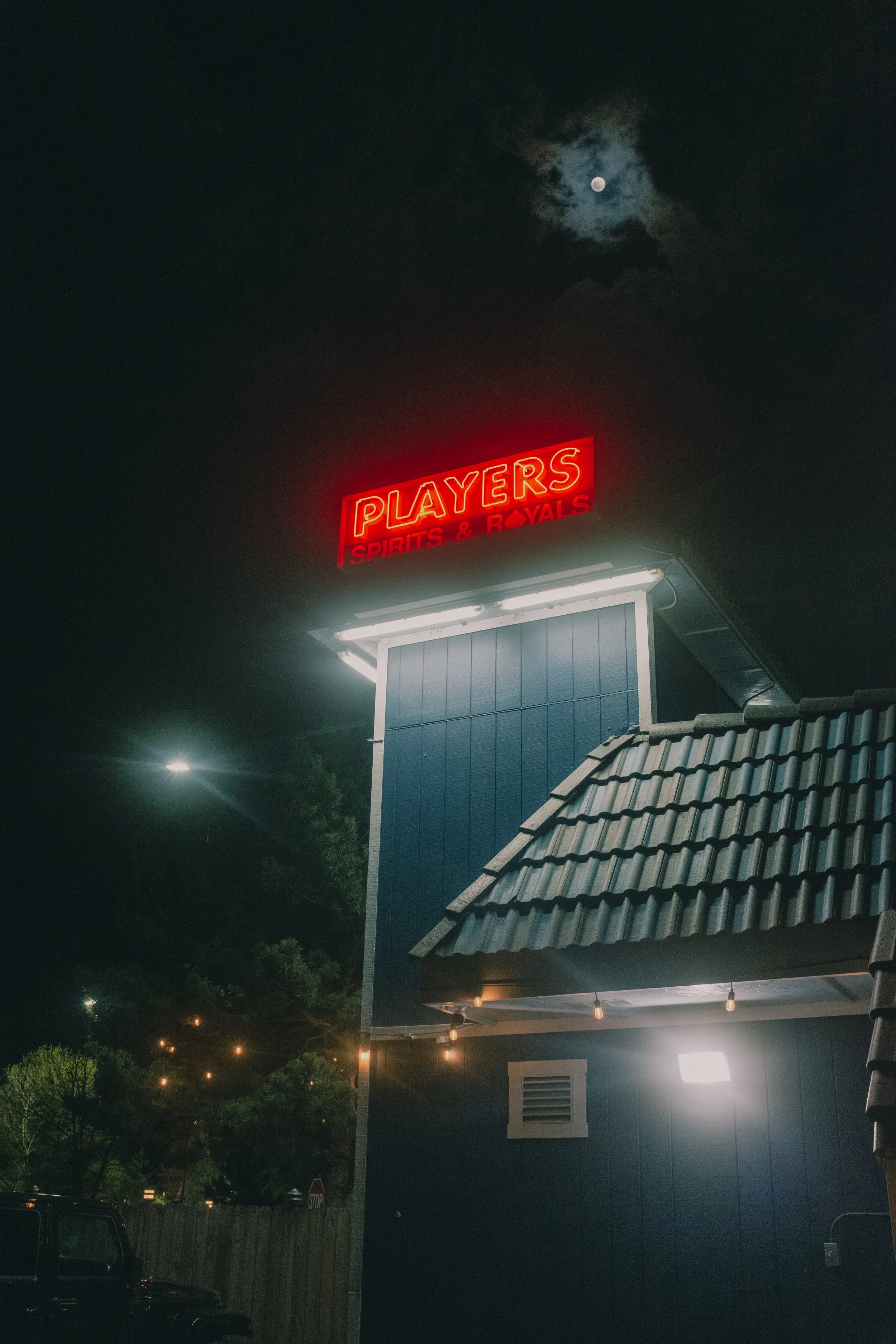 Nighttime scene with a blue building featuring a red neon sign that reads "PLAYERS SPIRITS & ROYAL" and a visible moon in the cloudy sky.