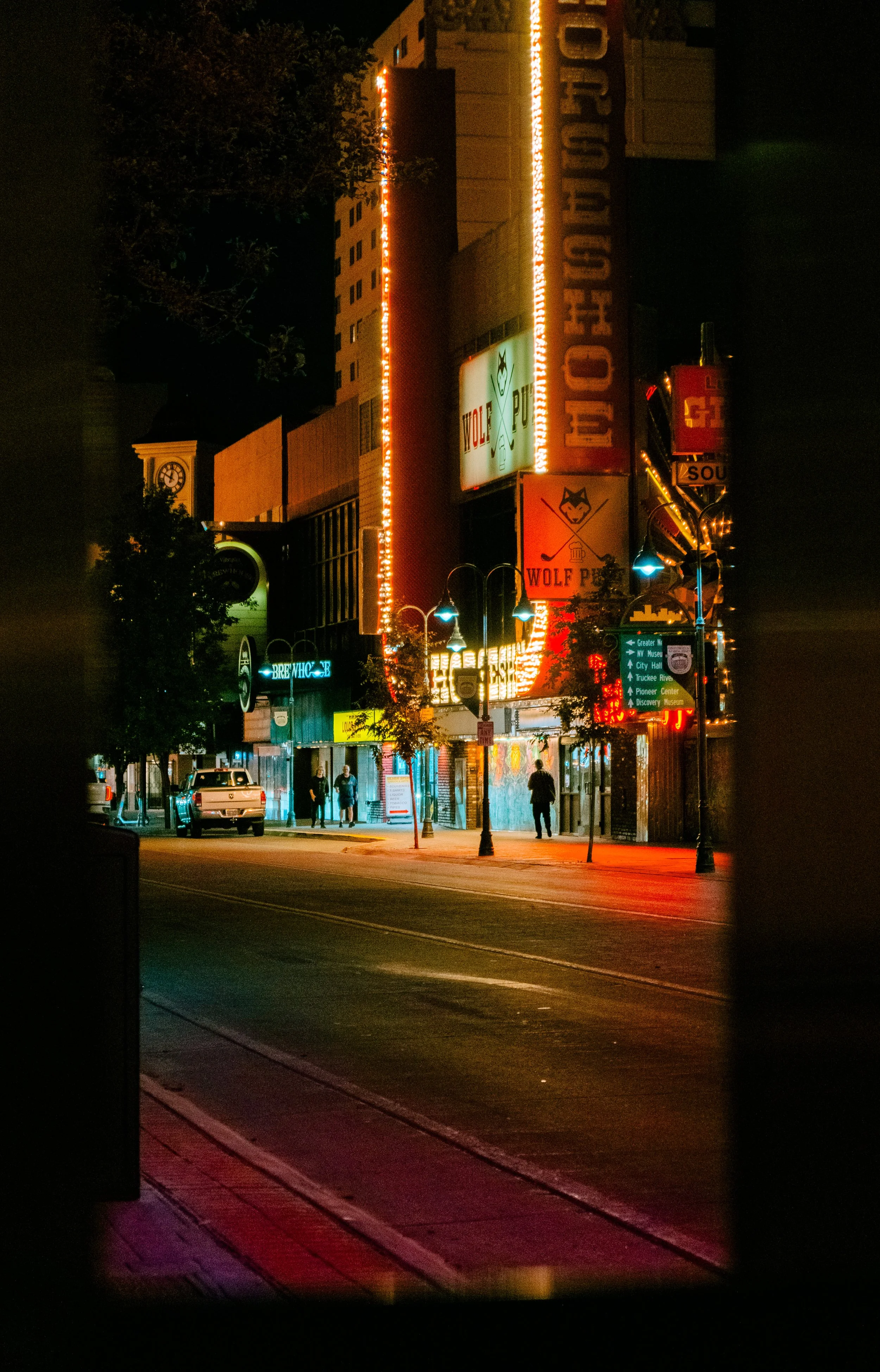 Nighttime city street view through a framed opening, illuminated signs and neon lights, people walking on the sidewalk, parked cars visible.