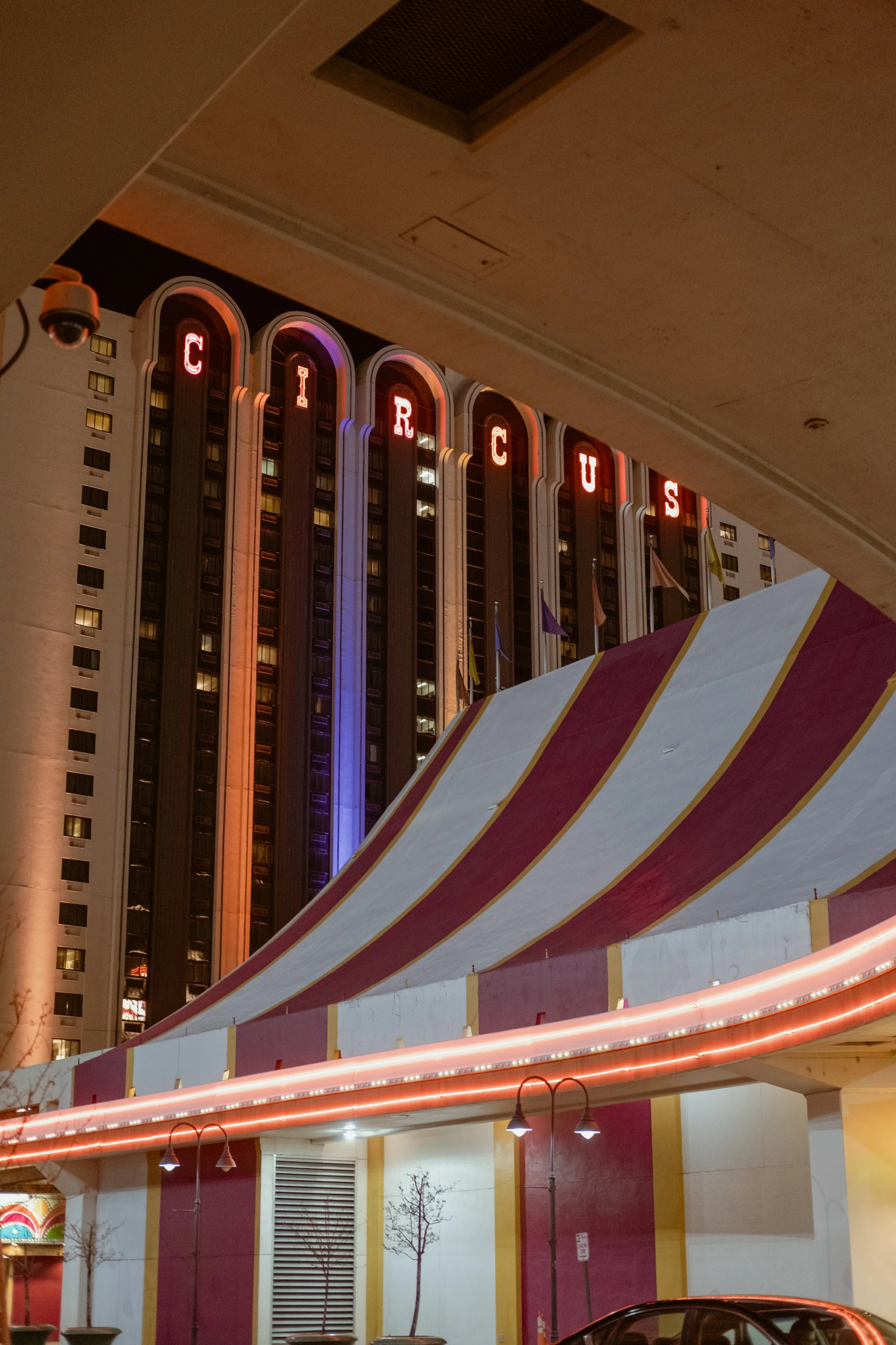 Night view of Circus Circus hotel and casino with illuminated vertical letters spelling 'CIRCUS' on the building, seen from beneath a canopy with red and white stripes.
