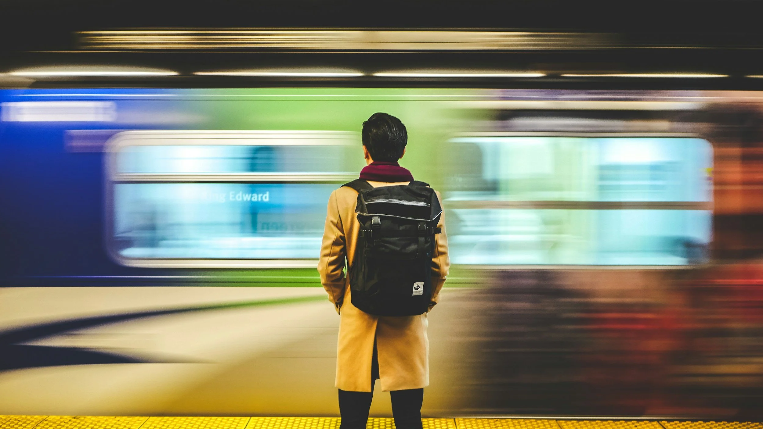 A person with a backpack standing on a subway platform as a train speeds by in a blur, with the person's back facing the camera.