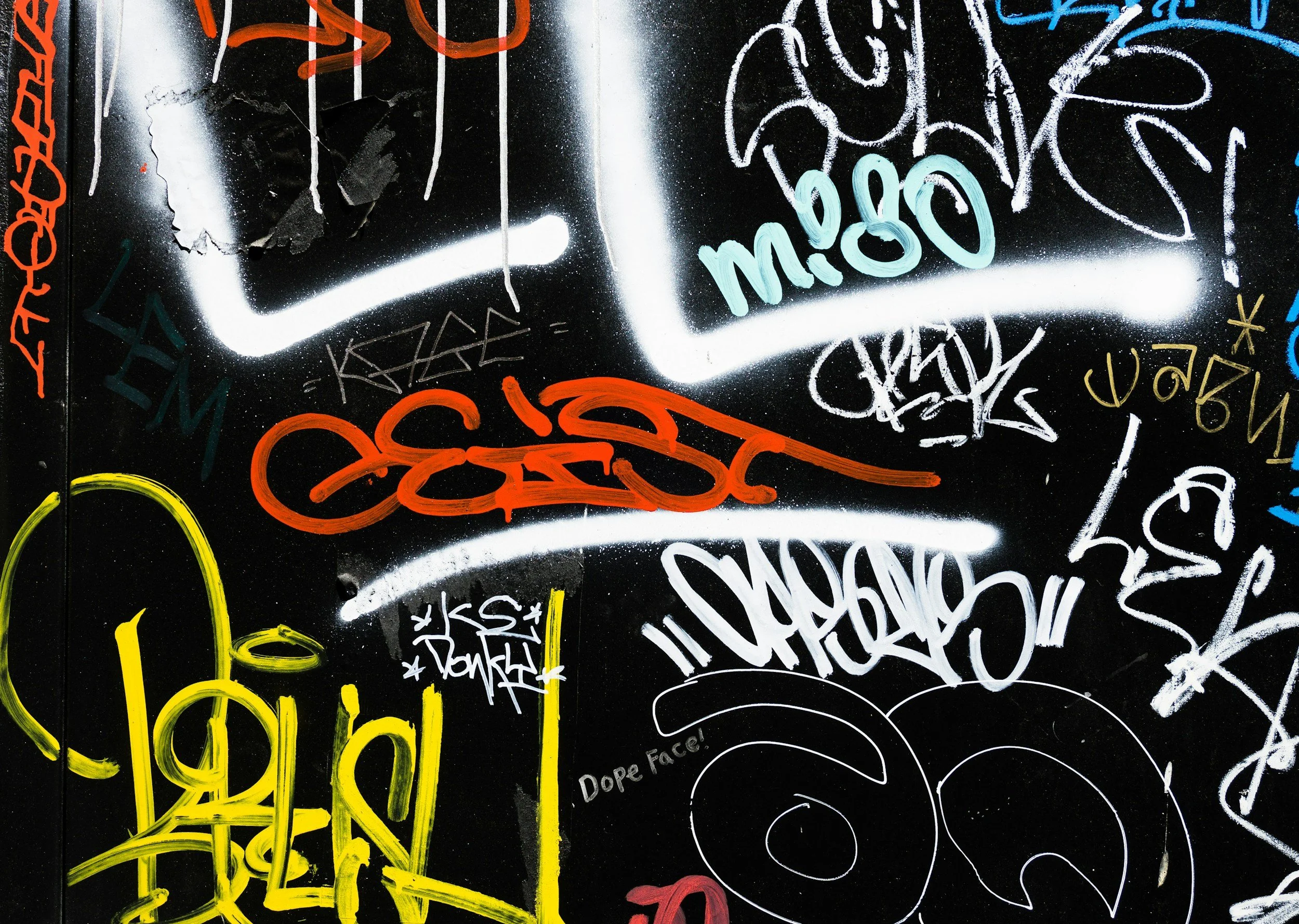 Graffiti wall with various colorful tags and writing, including yellow, red, blue, white, and orange spray-painted words and designs on a black background.
