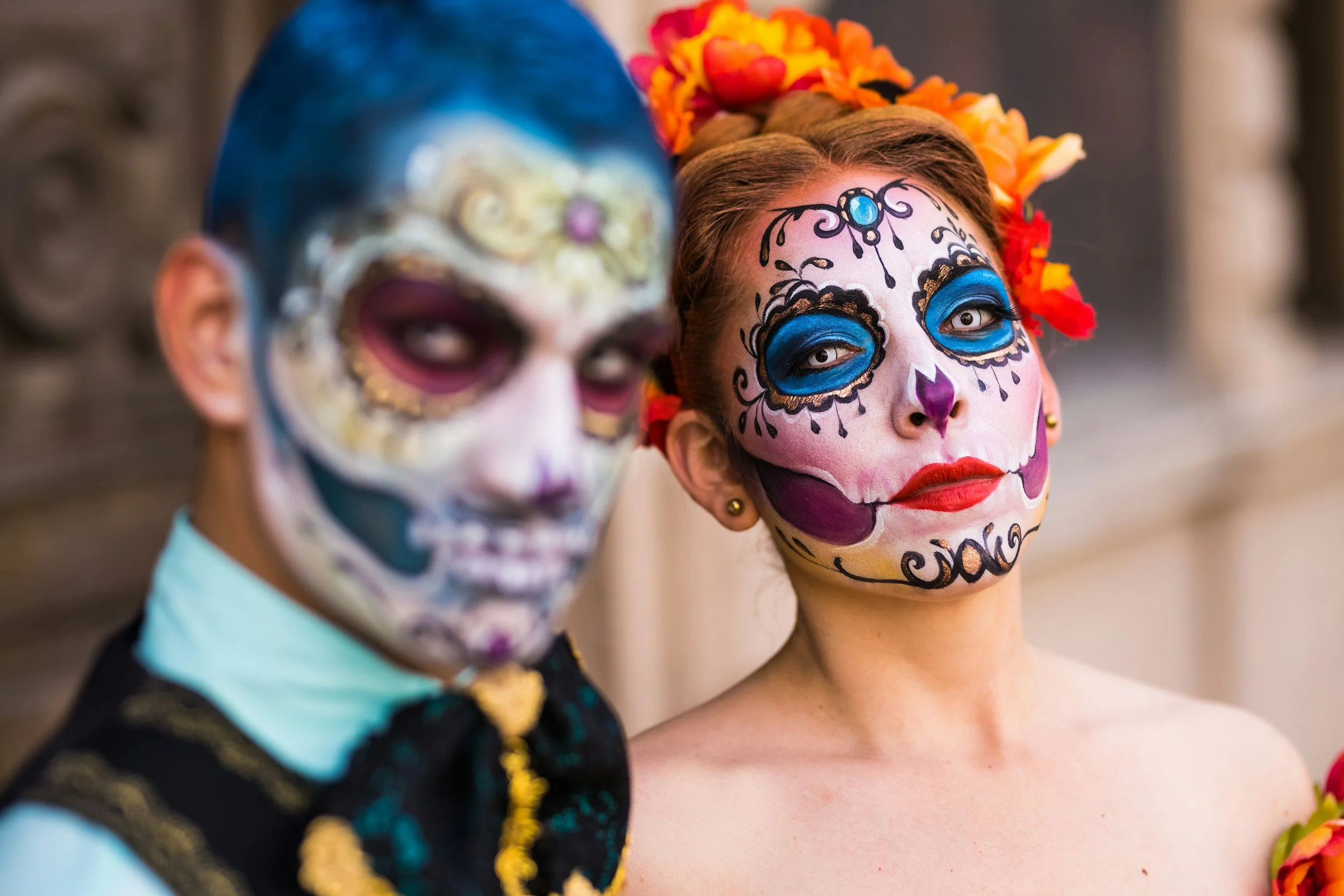 Two people with elaborate sugar skull makeup for Day of the Dead celebration, one woman wearing a flower crown and a shirtless man with a bow tie and vest.