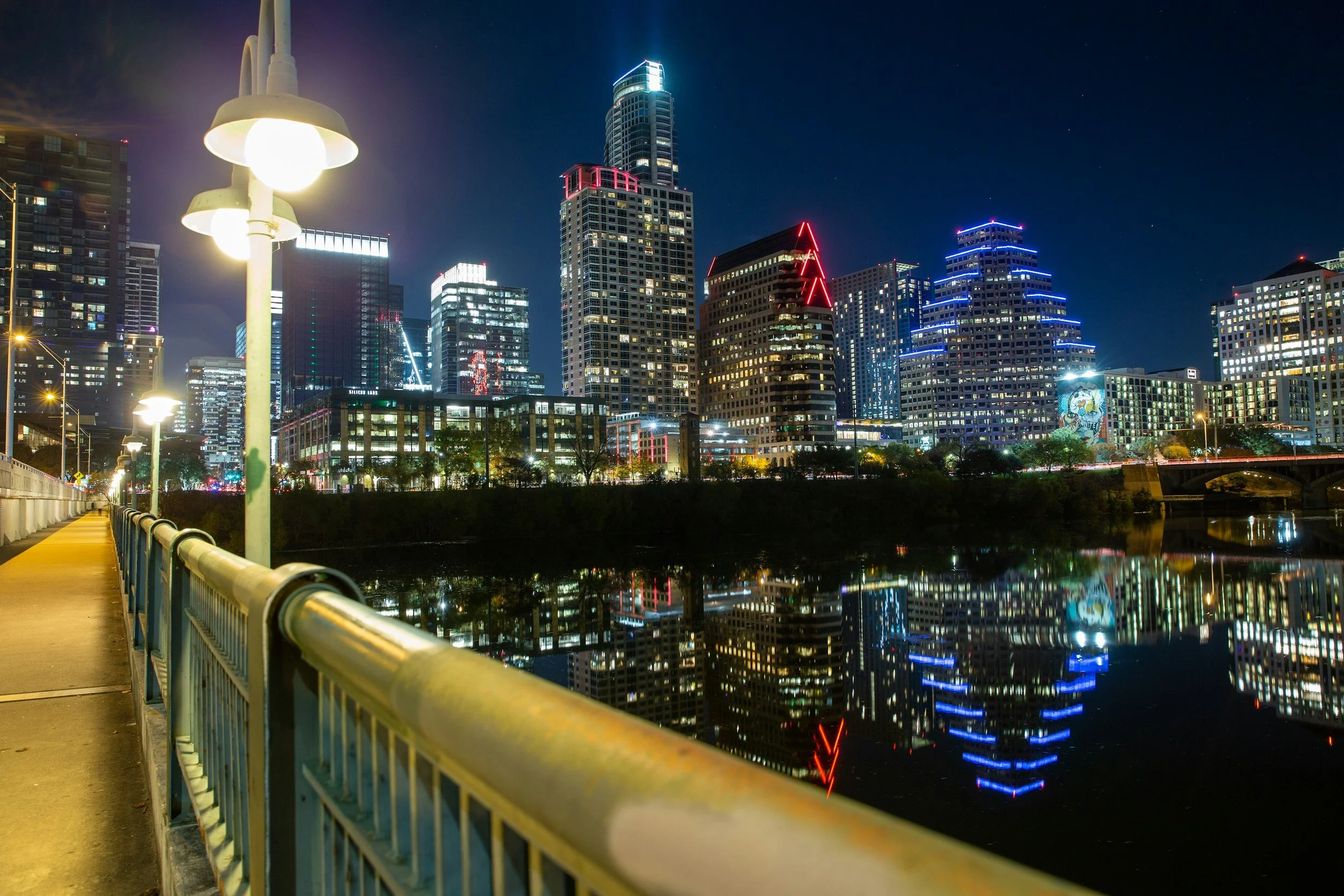 Nighttime cityscape of downtown with illuminated skyscrapers across a river, reflected in the water, viewed from a sidewalk with a fence and streetlights.