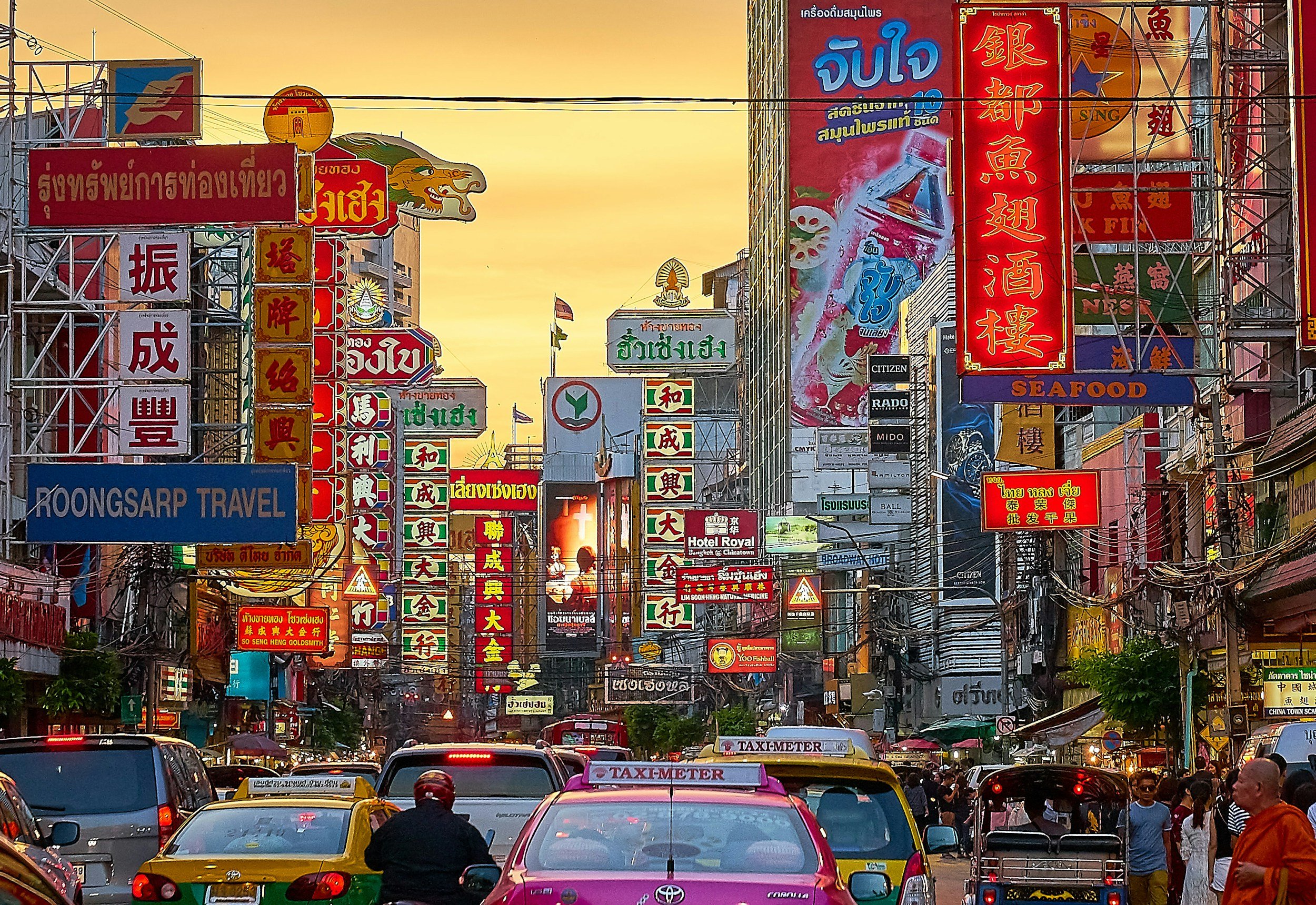 Busy street in Chinatown with numerous colorful signs in Chinese and Thai, taxis, pedestrians, and buildings under a sunset sky.