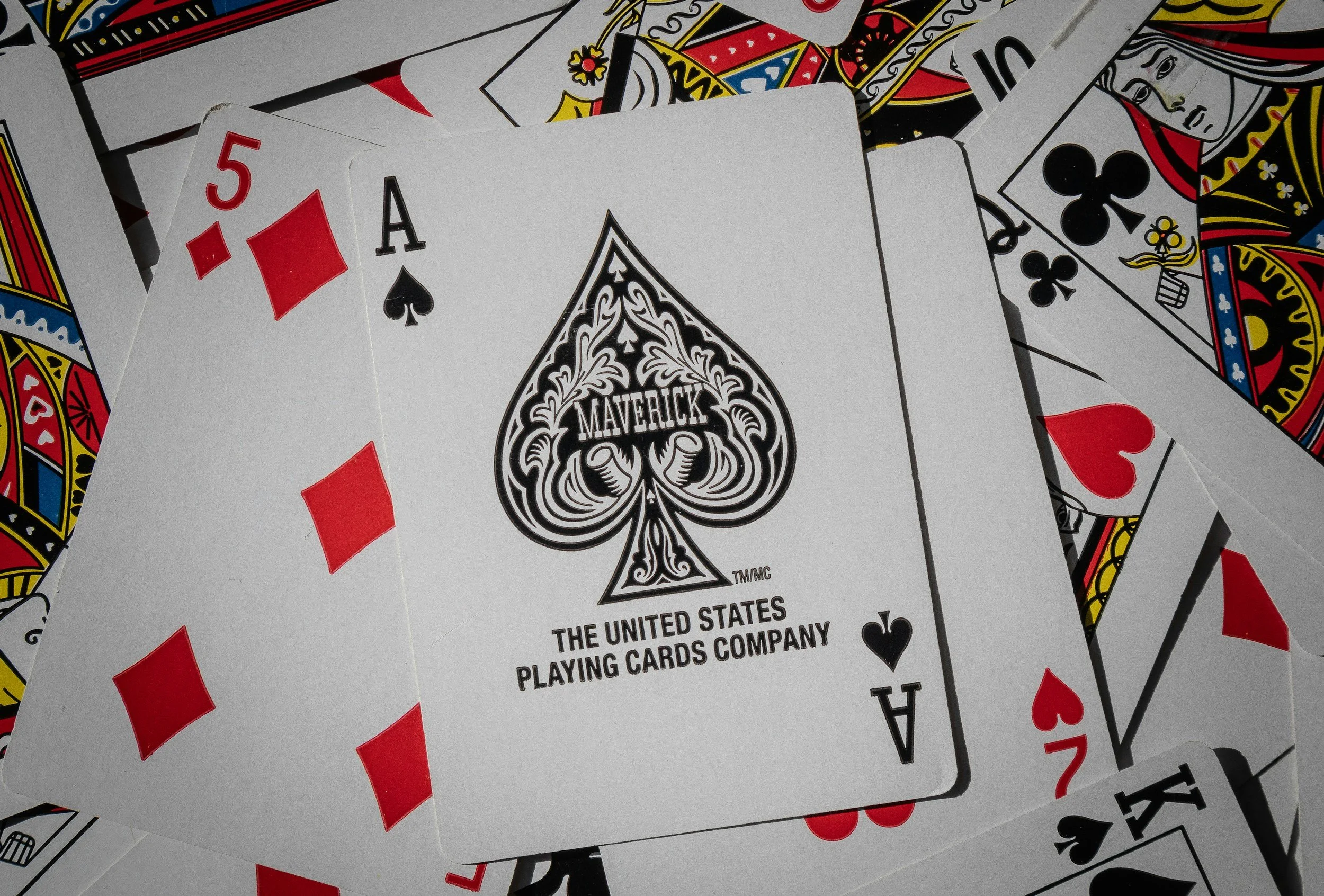 A deck of playing cards with the seven of hearts, ace of spades, five of diamonds, king of clubs, and other cards scattered around. The ace of spades features the Maverick logo and the text 'The United States Playing Cards Company.'