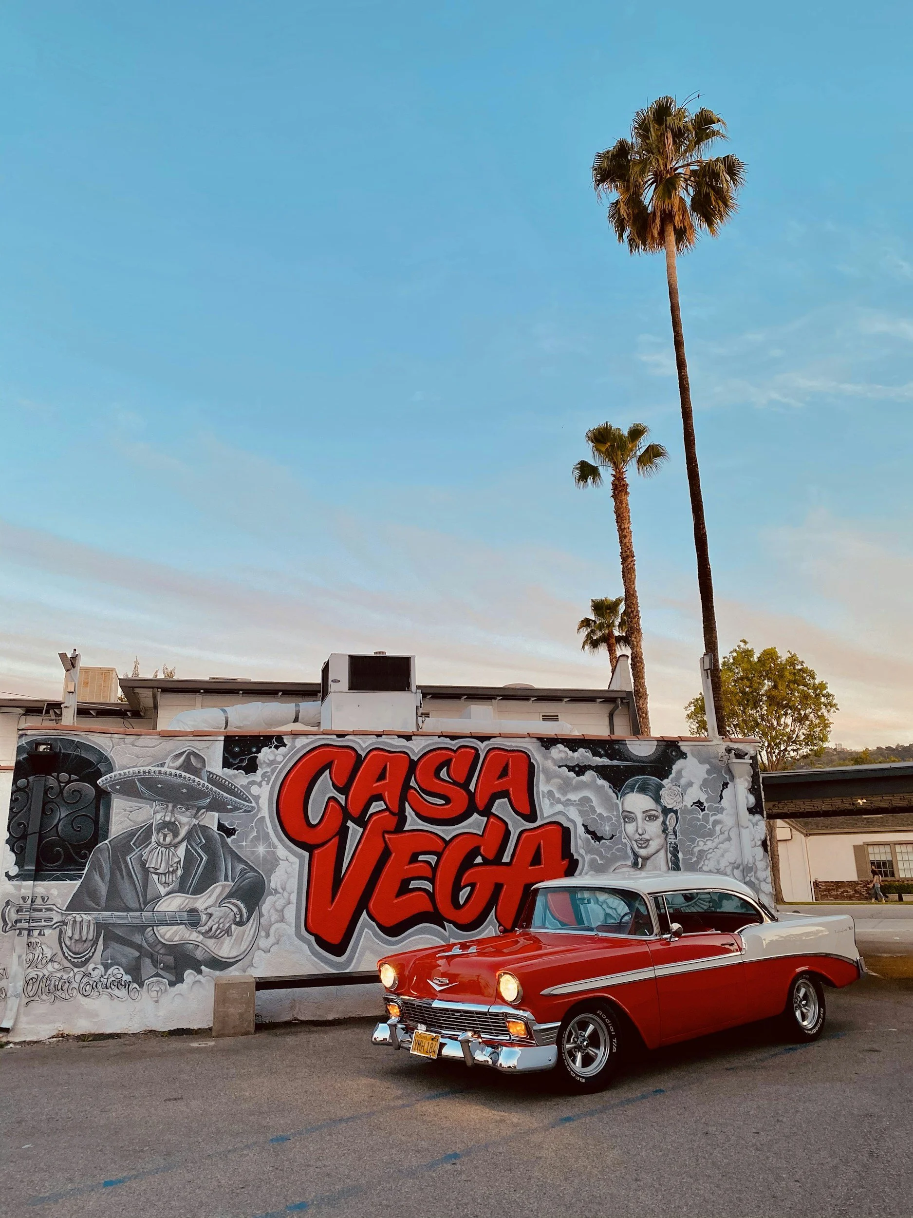 Red and white vintage car parked in front of a mural that reads "Casa Vega" with images of a musician with a guitar and a woman, and tall palm trees against a blue sky.