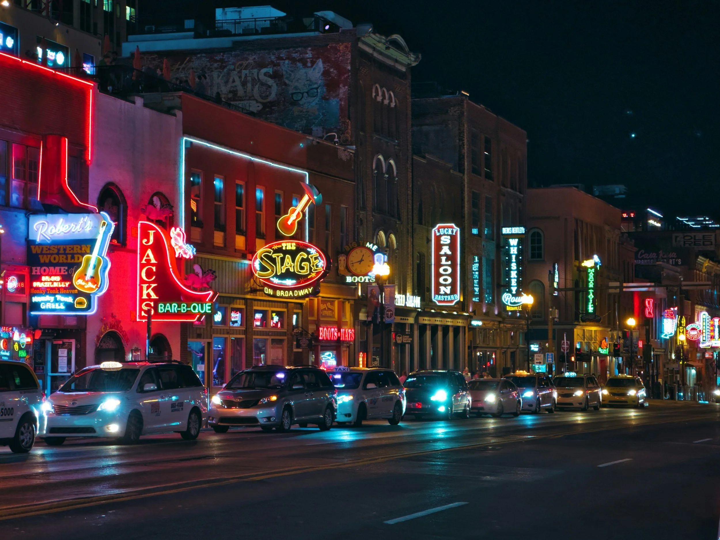 Night city street scene with neon signs for bars, restaurants, and entertainment venues, and parked cars along the curb.
