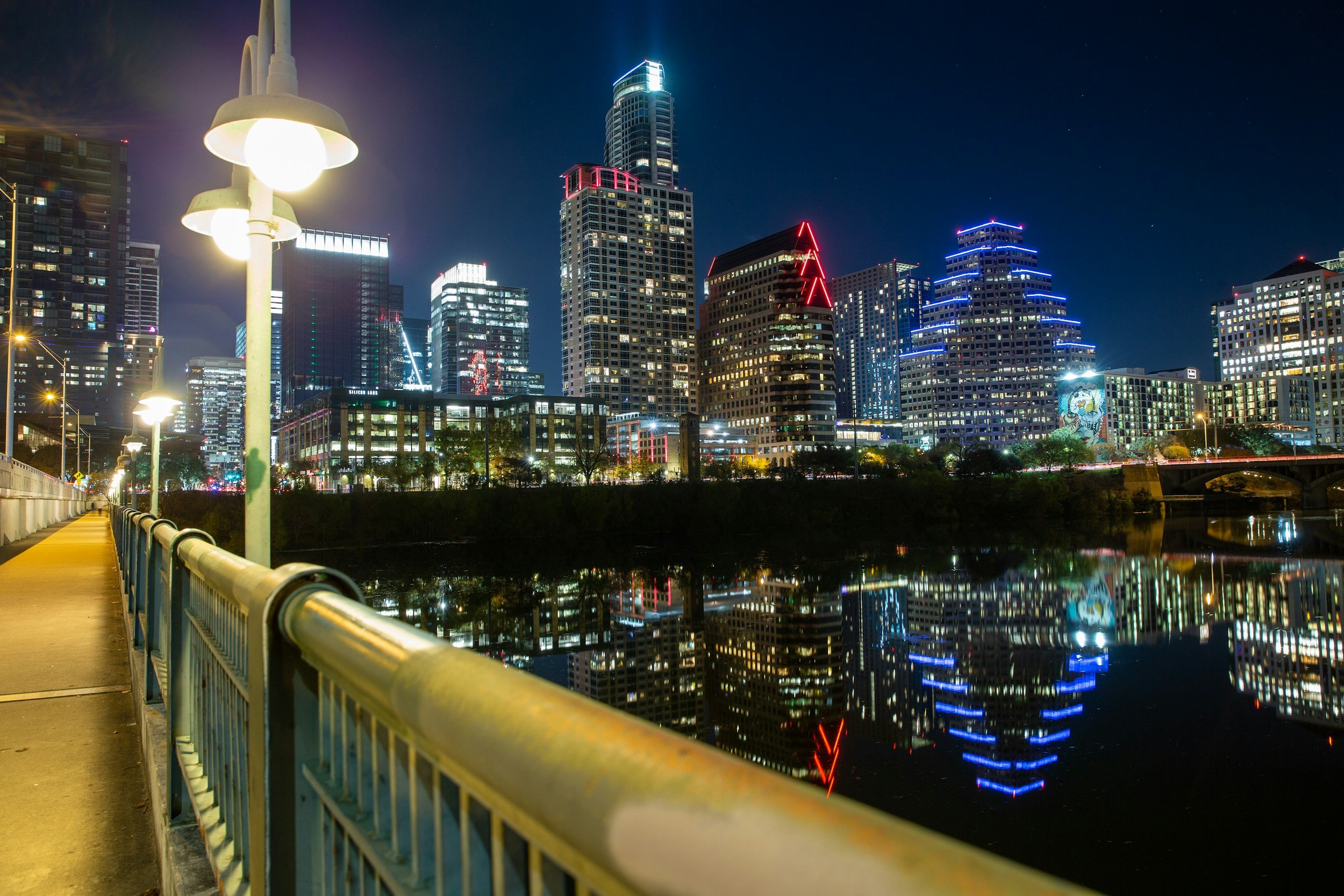 Nighttime cityscape with illuminated skyscrapers reflected in the river, seen from a riverside walkway with streetlights.