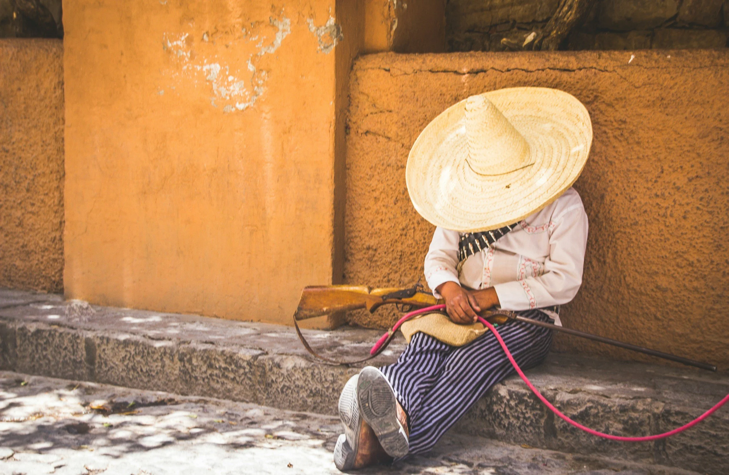 A person sitting on the sidewalk against a orange wall, wearing a large straw hat that covers their face, holding a musical instrument, and dressed in striped pants and a long-sleeved shirt.