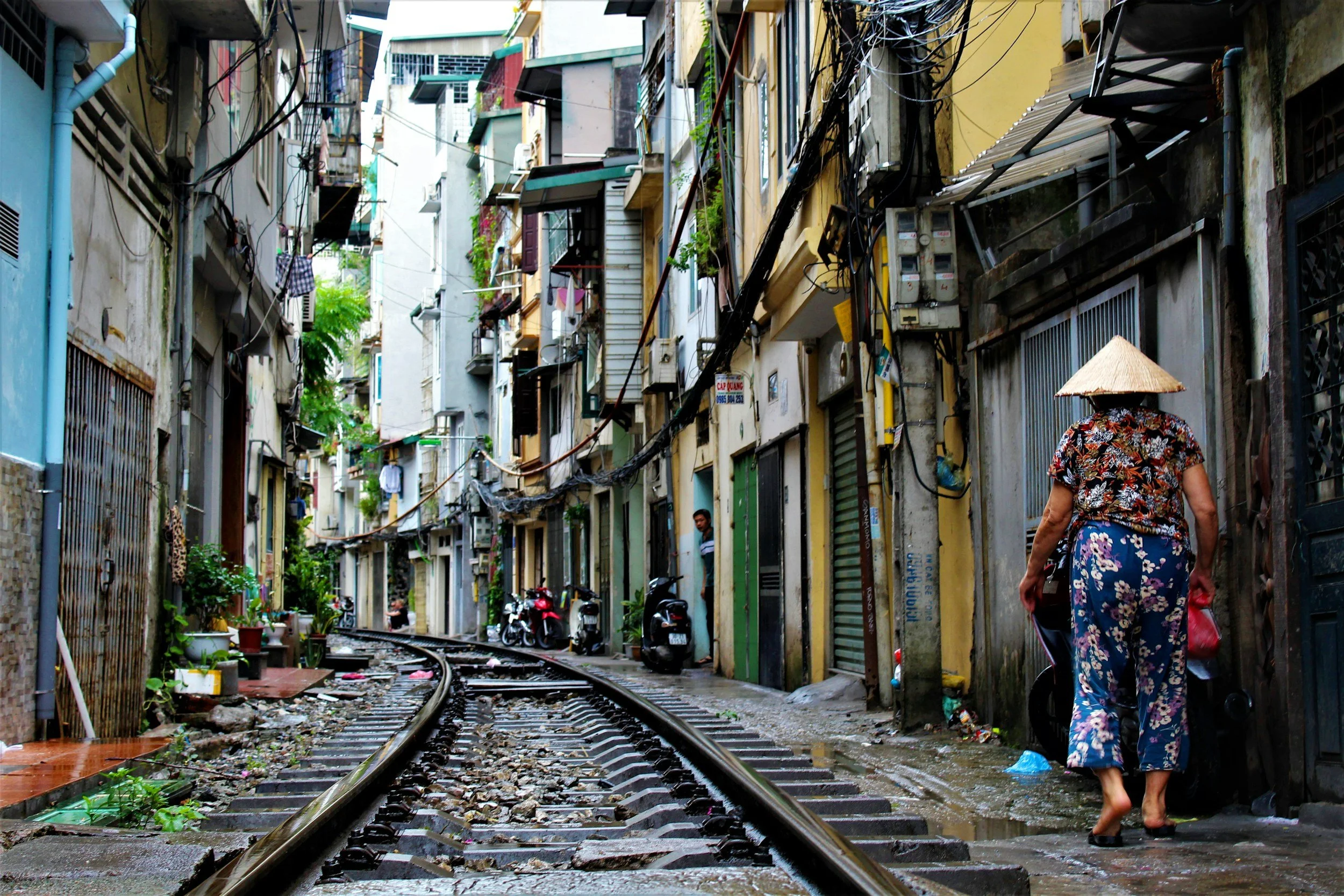 An urban alleyway with train tracks running through it, lined with multicolored apartment buildings, this scene shows an elderly woman in traditional clothing and a conical hat walking barefoot along the wet, muddy path. Motorcycles are parked on the