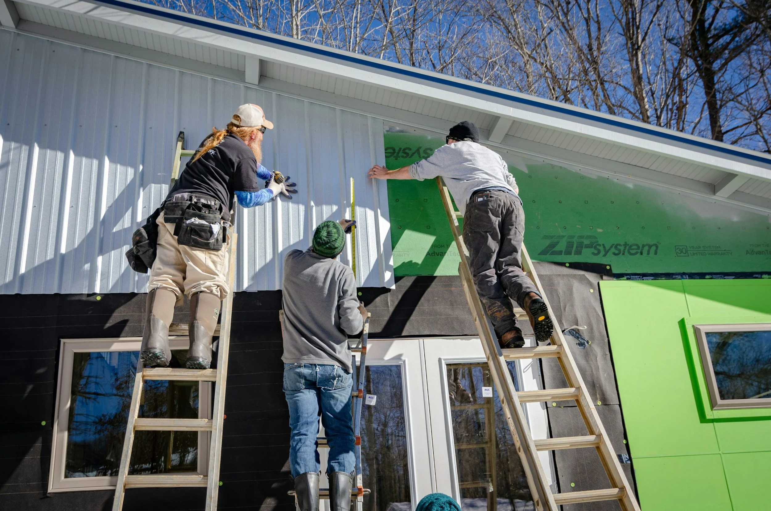 Four construction workers installing siding or exterior wall material on a building. Two are on ladders and two on the ground, working together to attach green and white panels to the building's exterior.
