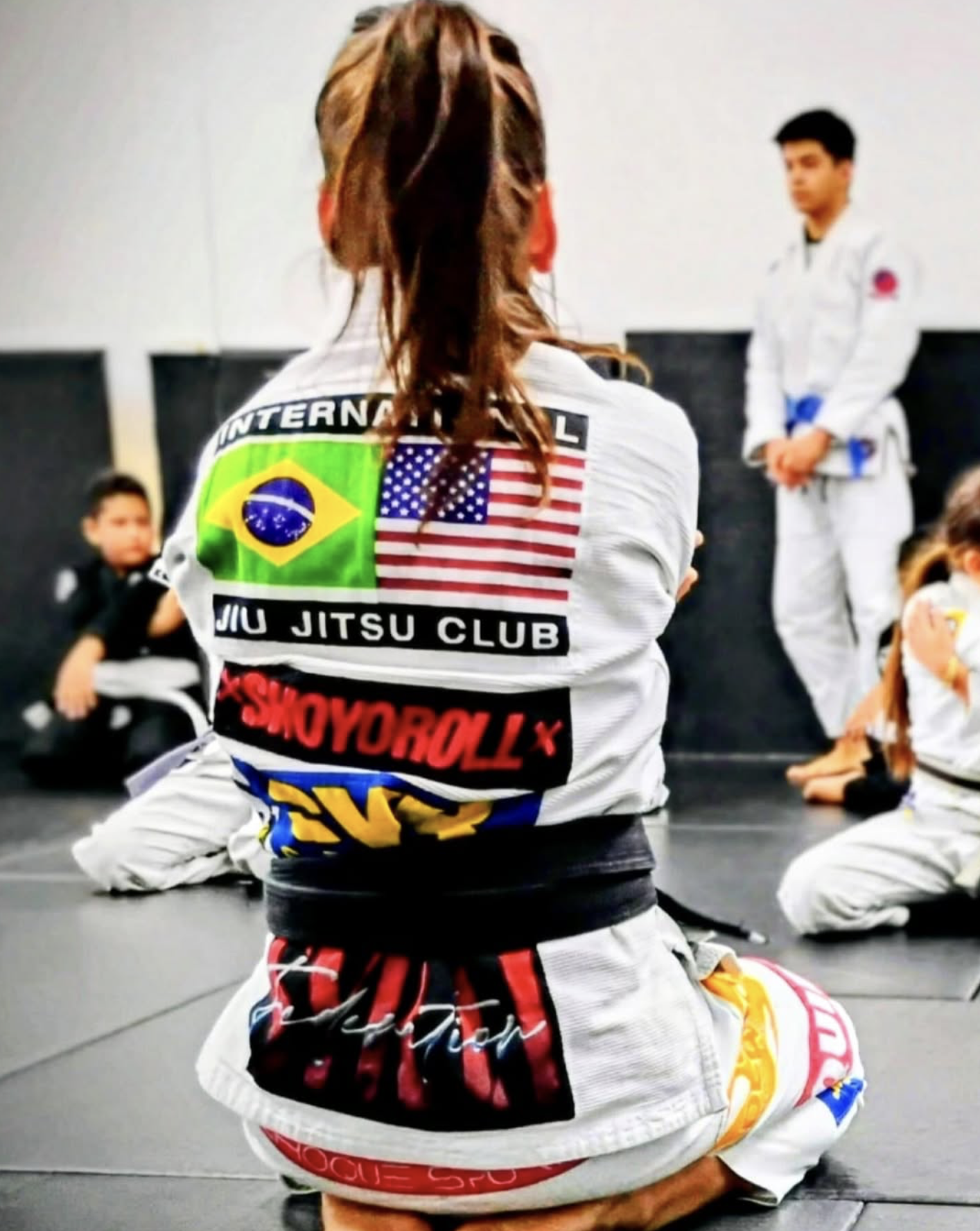 Brazilian woman in Brazilian Jiu Jitsu gi kneeling on the mat, with her back to the camera, facing a male instructor and other students in Gi inside a martial arts dojo.